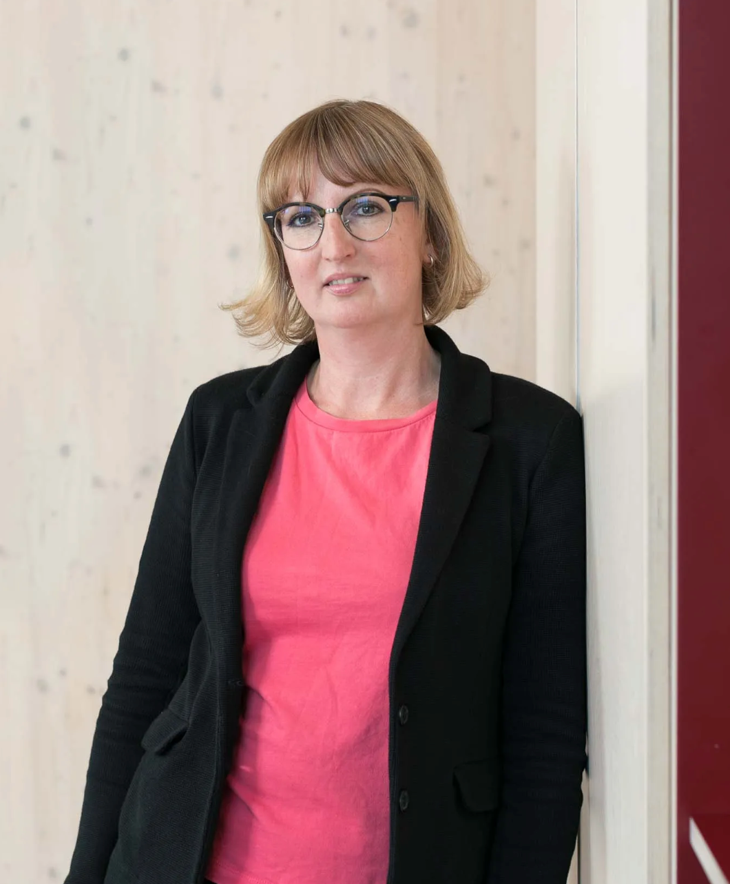Woman in black blazer and pink top with glasses poses confidently against a light wooden wall.