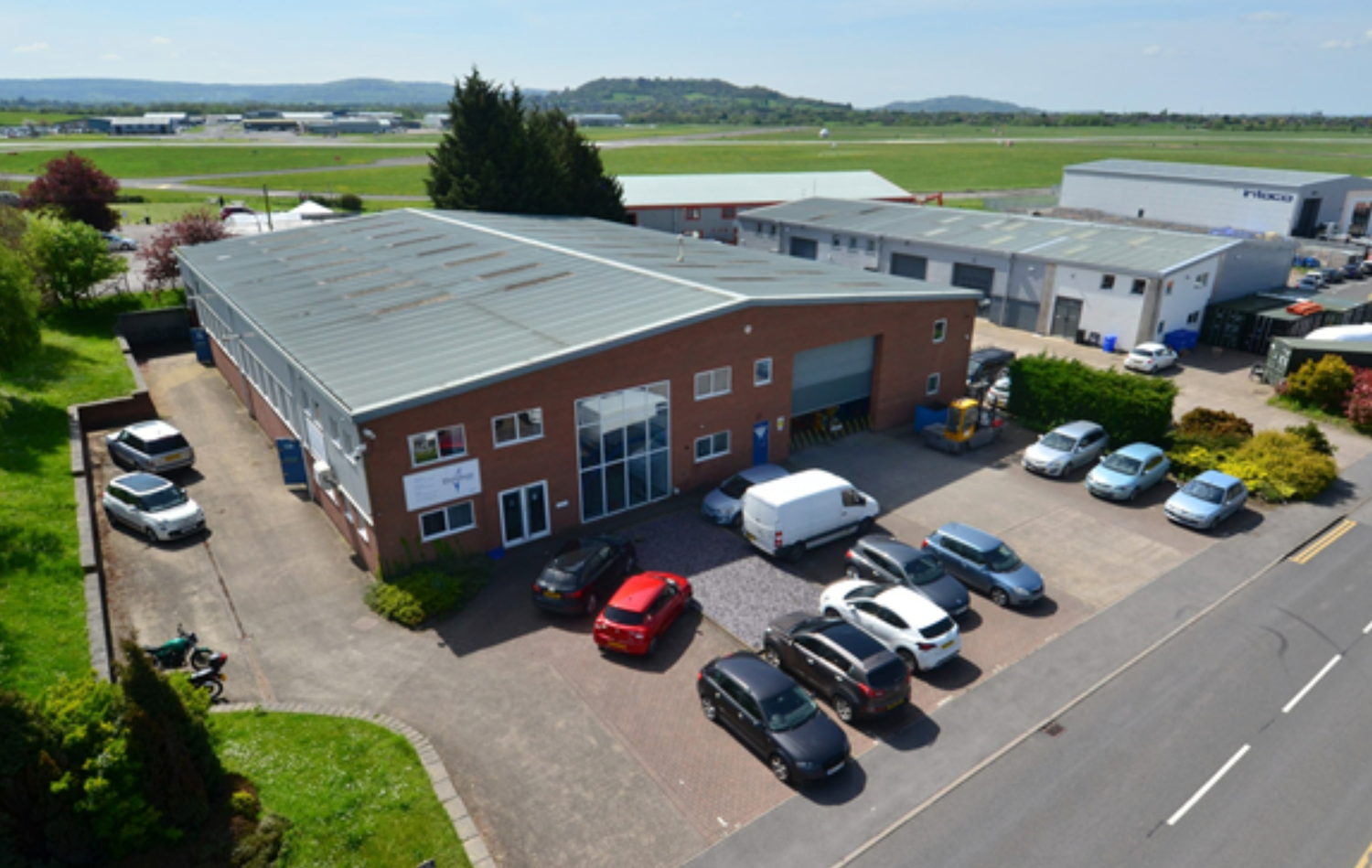 Aerial view of a modern industrial building with parking lot, green fields, and runway in background on a sunny day.