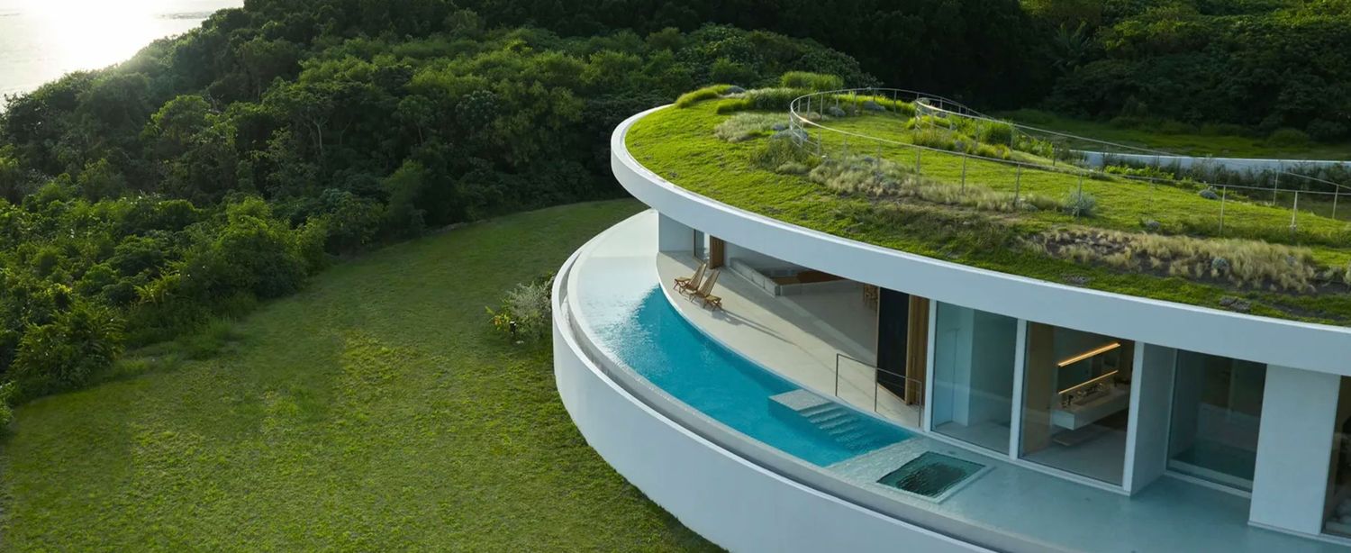 Aerial view of a modern curved villa with a green roof garden and infinity pool nestled into a lush hillside.