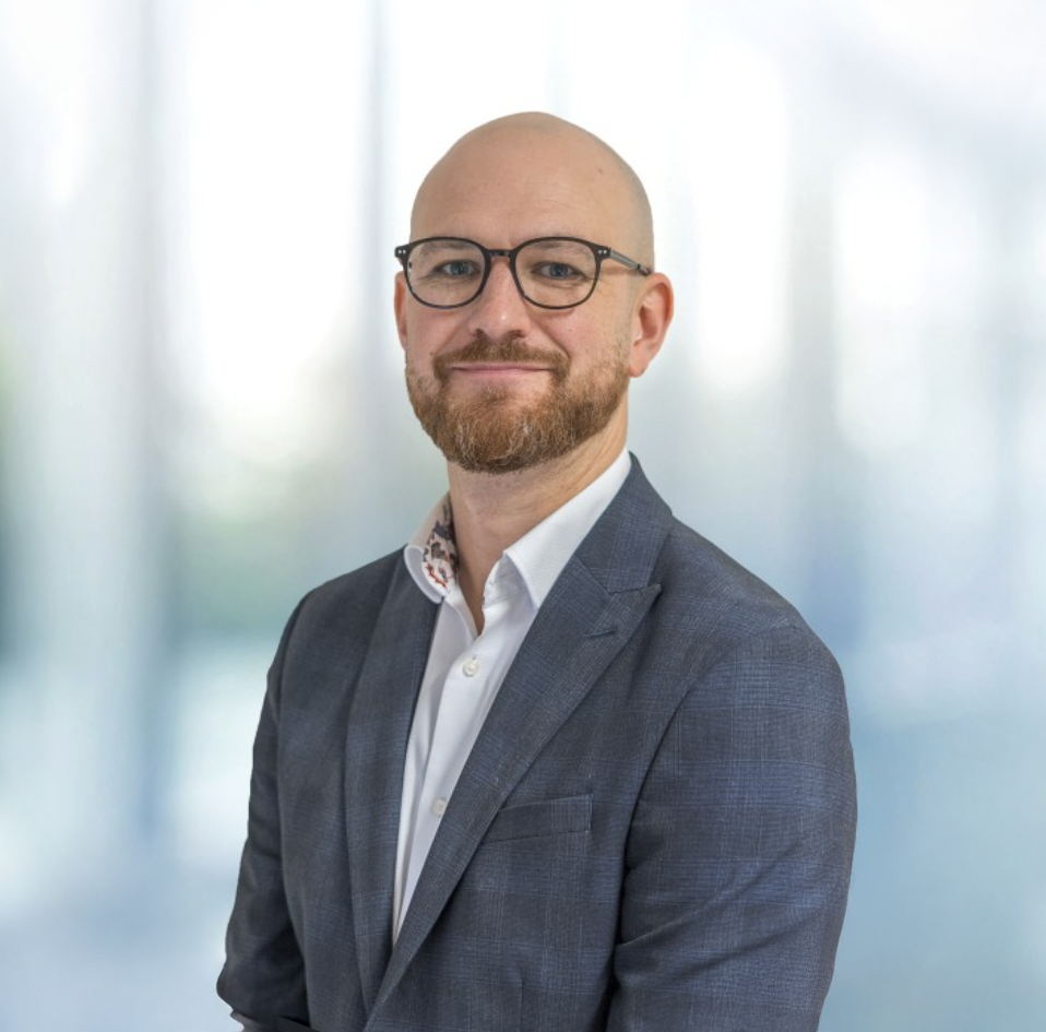 Bald professional man with glasses and beard wearing a blue blazer smiling in a modern office setting.