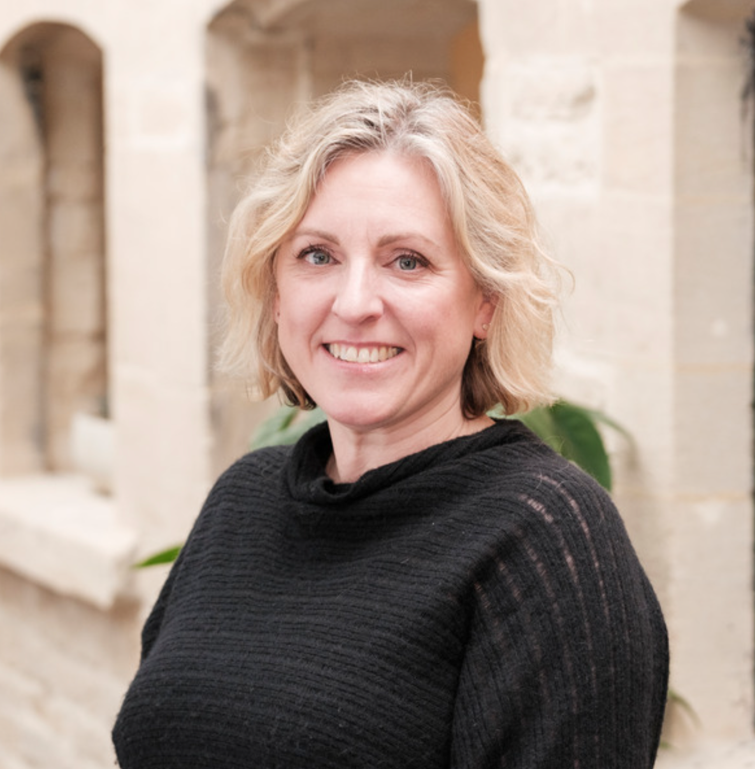Smiling blonde woman in black turtleneck sweater poses outdoors near stone architecture.