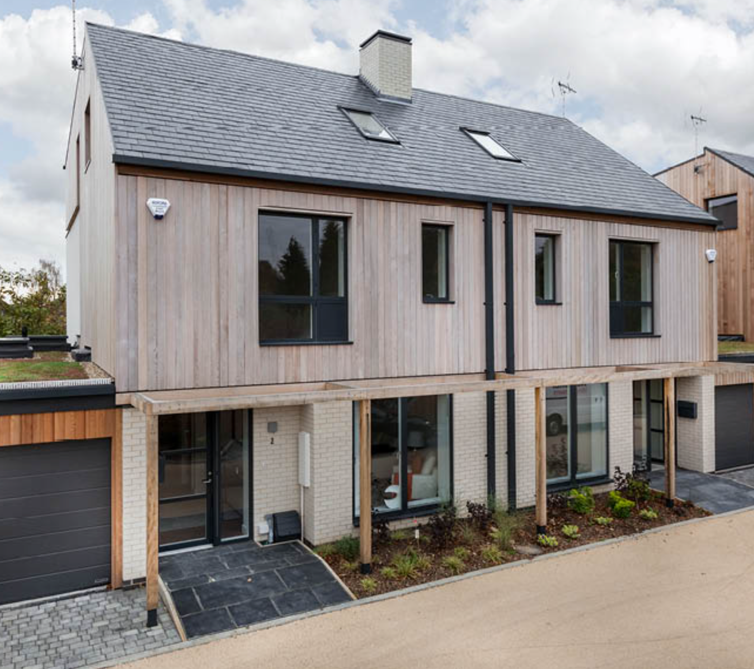 Modern semi-detached house with timber cladding, slate roof, large windows, and a garage in a residential setting.