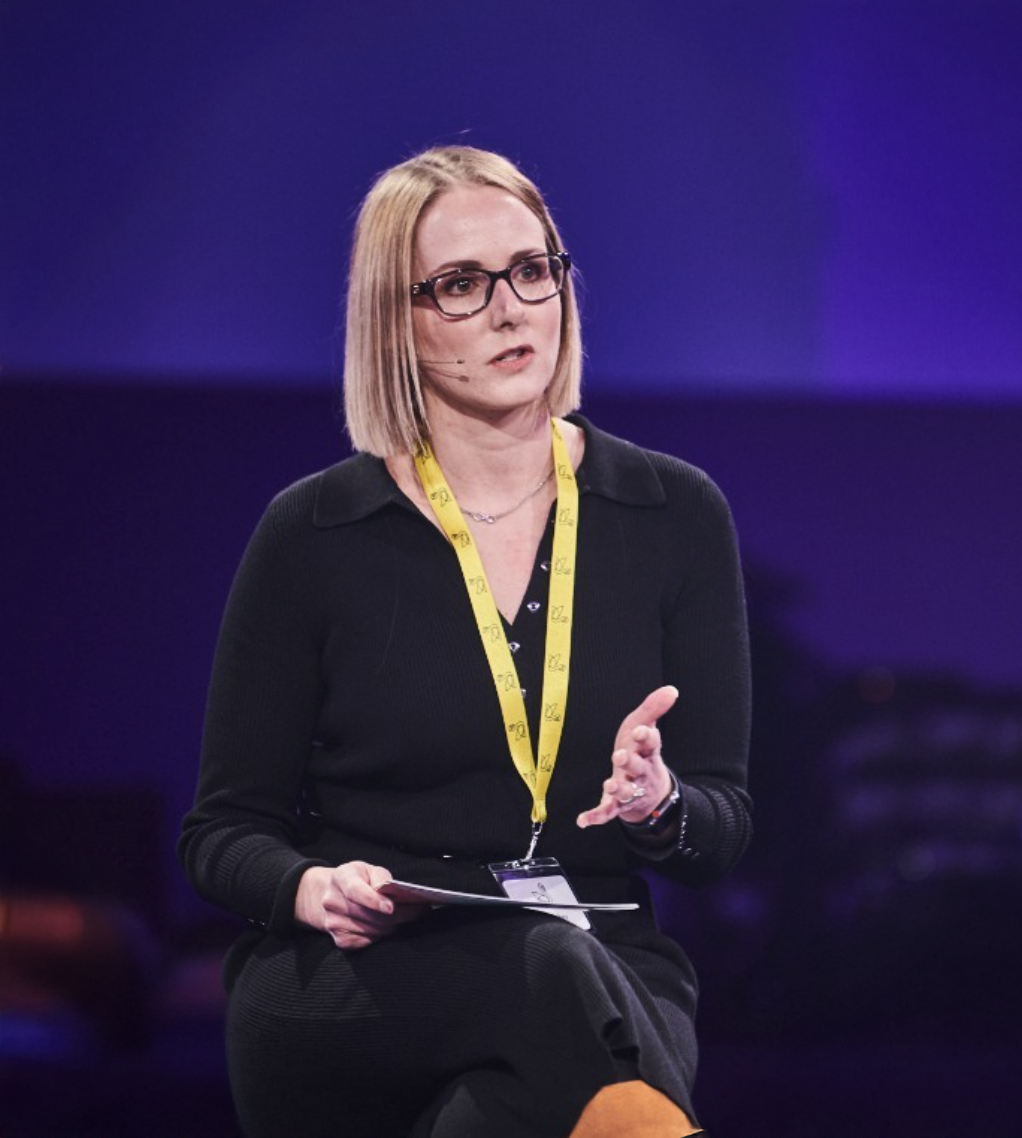 Blonde woman with glasses speaking at a conference, wearing black outfit and yellow lanyard on stage.