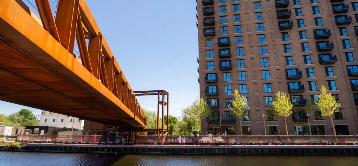 Rust-colored steel bridge spans a canal beside a modern brick high-rise building on a sunny day.