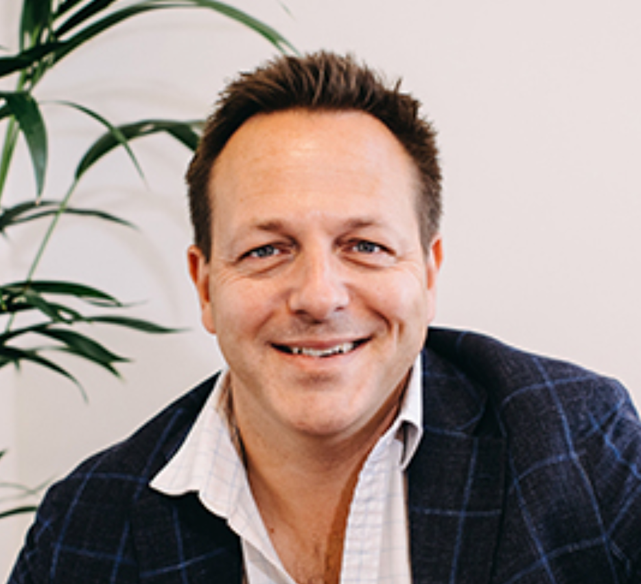 Smiling middle-aged man in a blue blazer and white shirt posing indoors near a tropical plant.