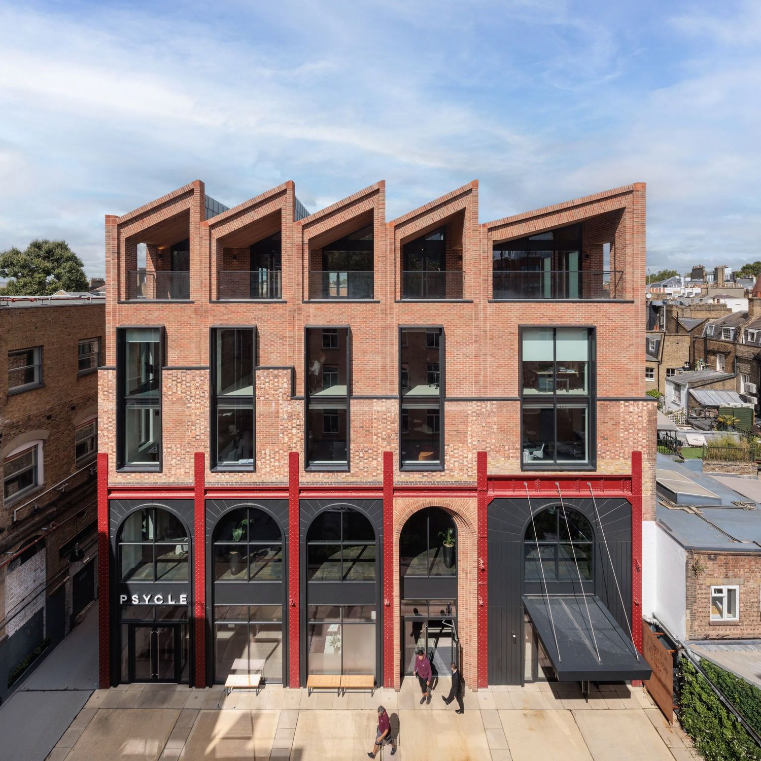 Modern red brick commercial building with arched ground floor windows and sawtooth roofline under blue sky.