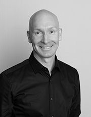 Professional black and white headshot of a smiling bald man in a dark button-up shirt against a light background.