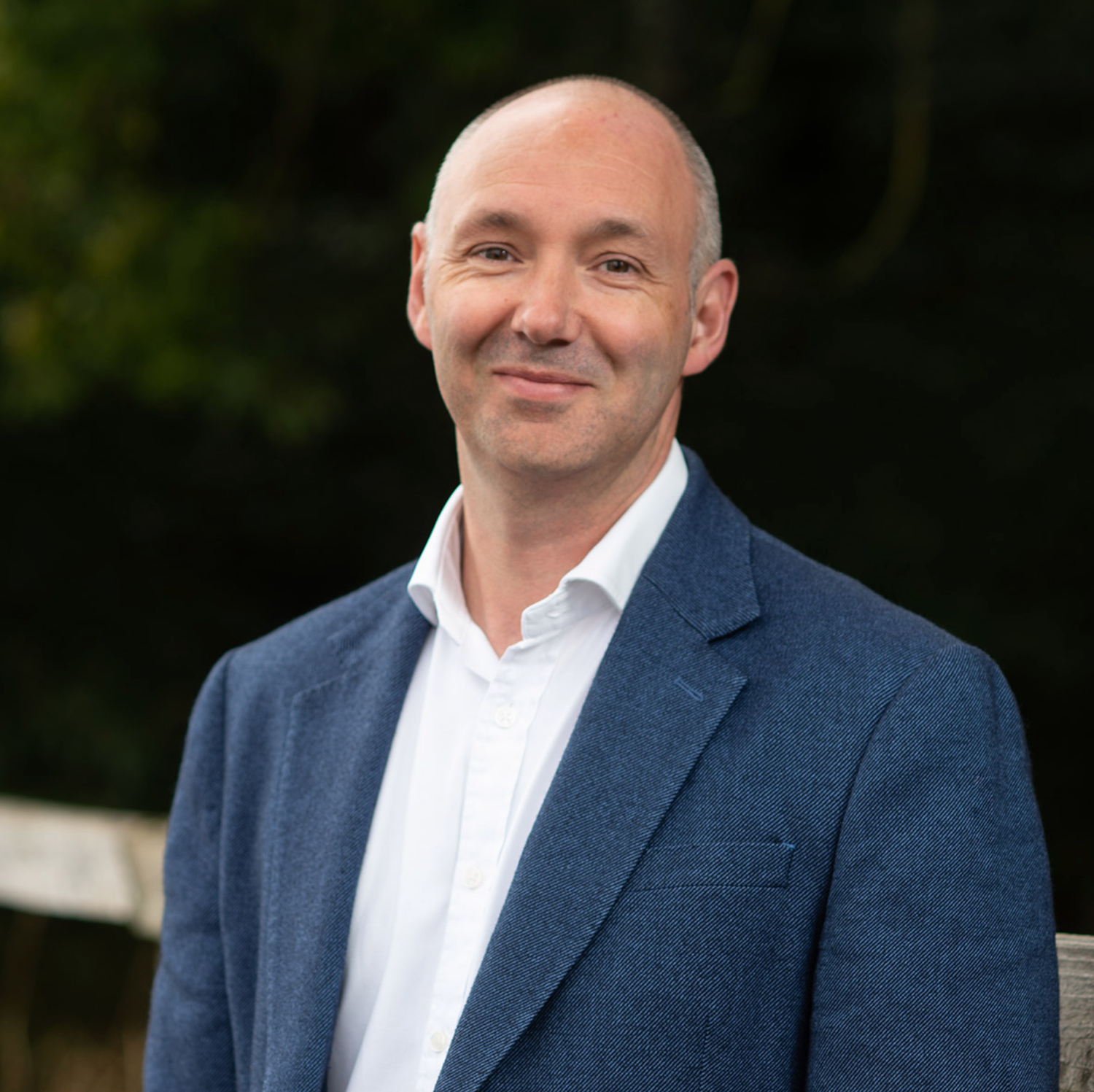 Professional man in blue blazer smiling outdoors with green foliage background, business portrait photo.