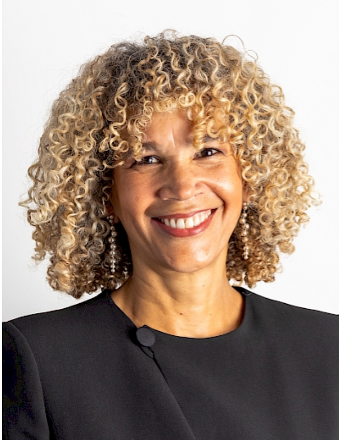 Professional headshot of a smiling woman with curly blonde hair wearing a black blazer against a white background.