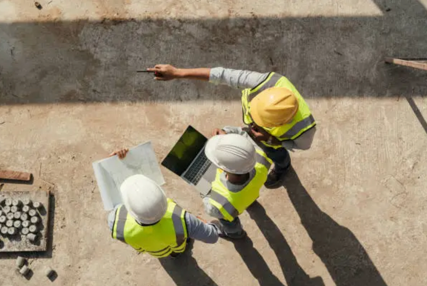 Three construction workers in safety vests and hard hats review plans on a laptop at a job site.