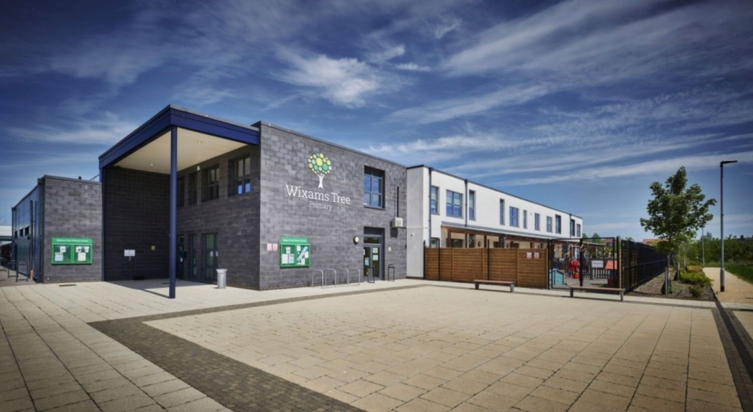 Modern school building with dark brick facade, covered entrance, and spacious paved courtyard under a blue sky.