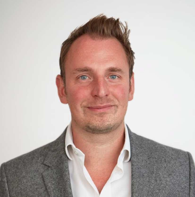Professional headshot of a smiling man with blue eyes and brown hair wearing a gray blazer and white shirt.