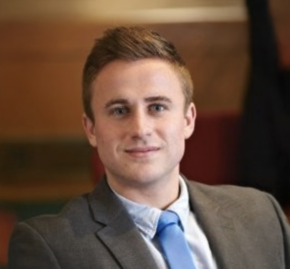 Young professional man in gray suit and blue tie smiling confidently in a formal indoor setting.