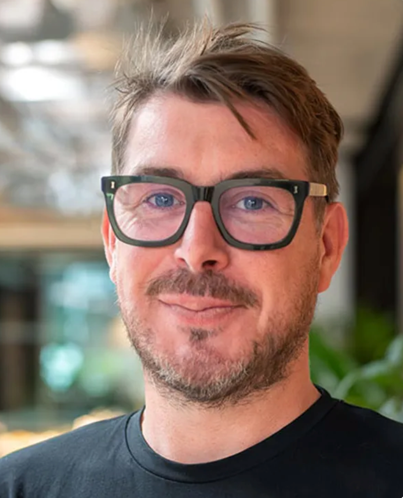 Man with dark-rimmed glasses and short brown hair smiling, wearing a black t-shirt in a modern indoor setting.