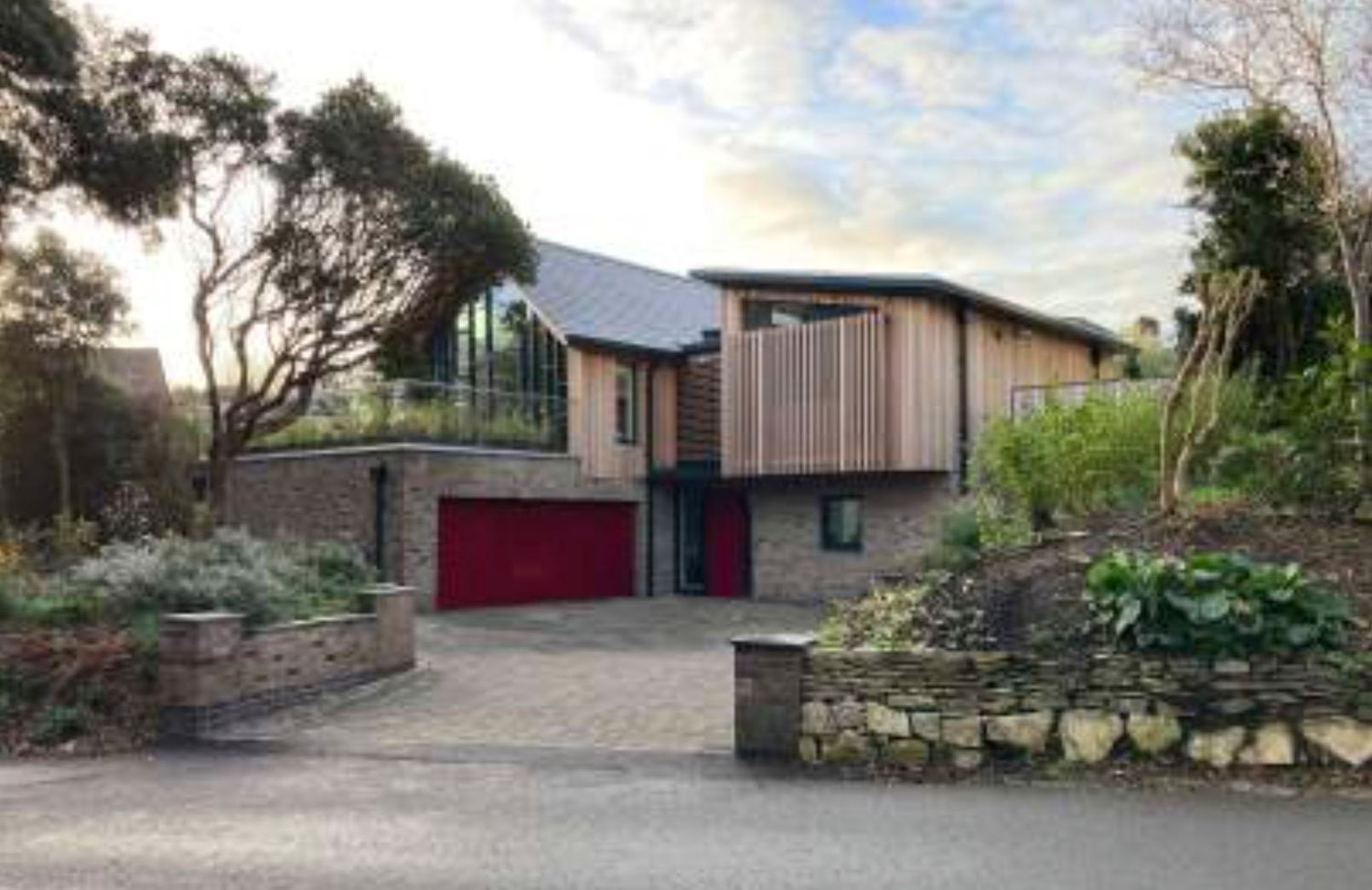 Modern brick home with red garage door, timber cladding, large glass windows, and landscaped garden on a curved driveway.