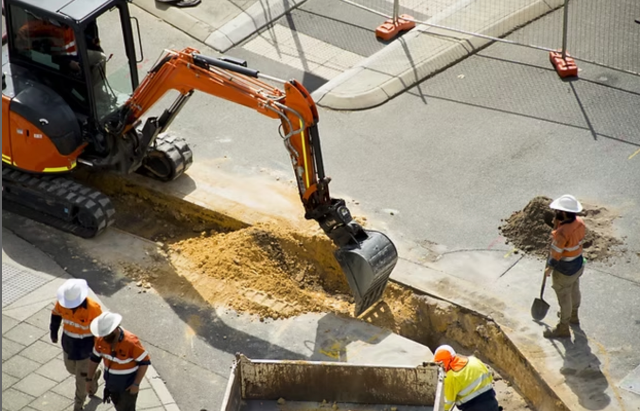 Orange excavator digging at a construction site with workers in high-visibility vests and hard hats supervising.