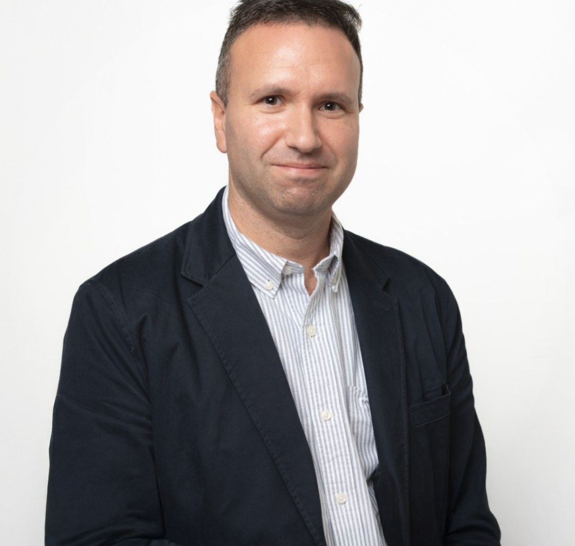 Professional man in dark blazer and striped shirt smiling against white background.