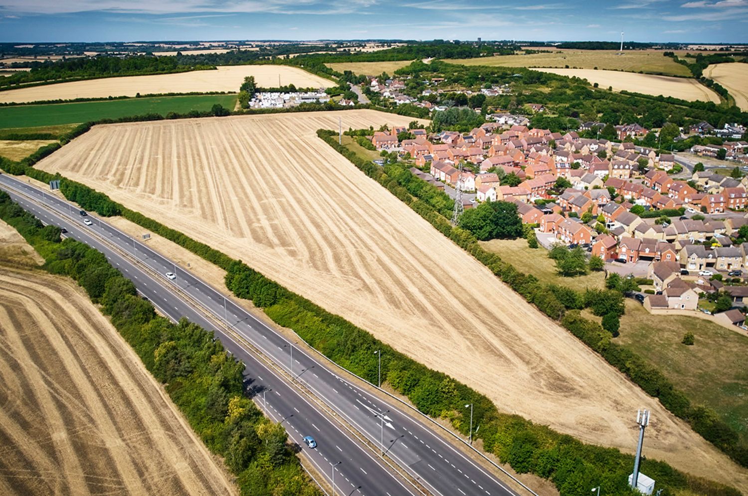 Aerial view of a harvested wheat field beside a dual carriageway road next to a residential village in rural England.