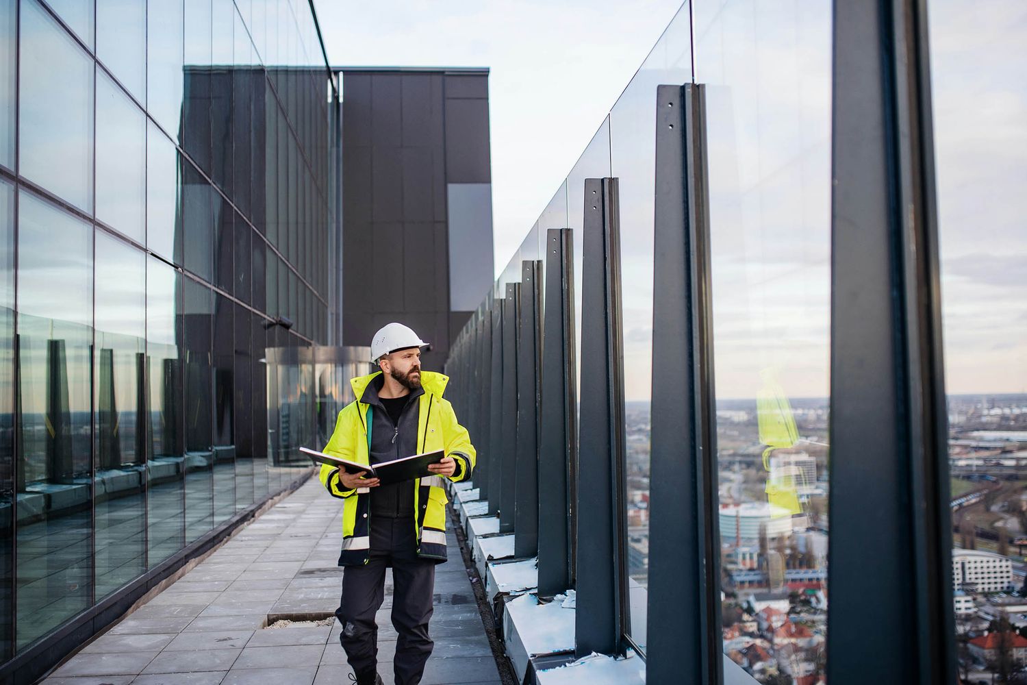 Construction worker in hi-vis jacket and hard hat inspecting a high-rise building exterior on a rooftop terrace.