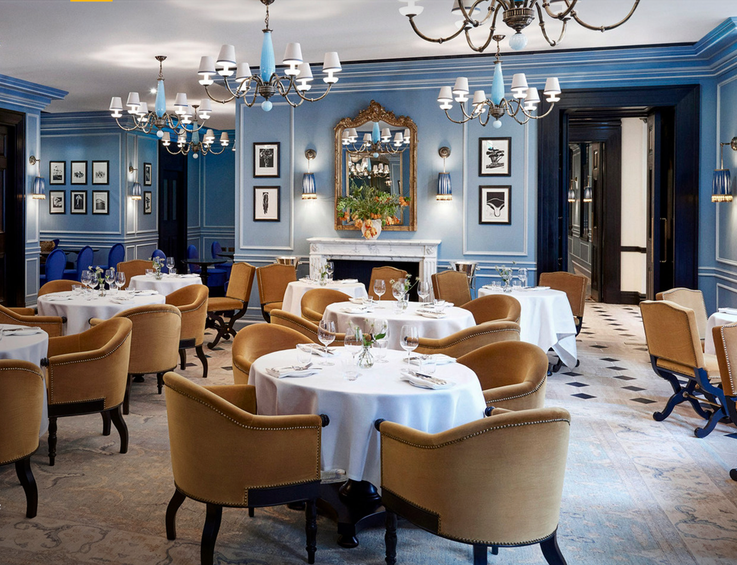 Elegant blue-walled fine dining restaurant with gold velvet chairs, white tablecloths, chandeliers, and a marble fireplace.