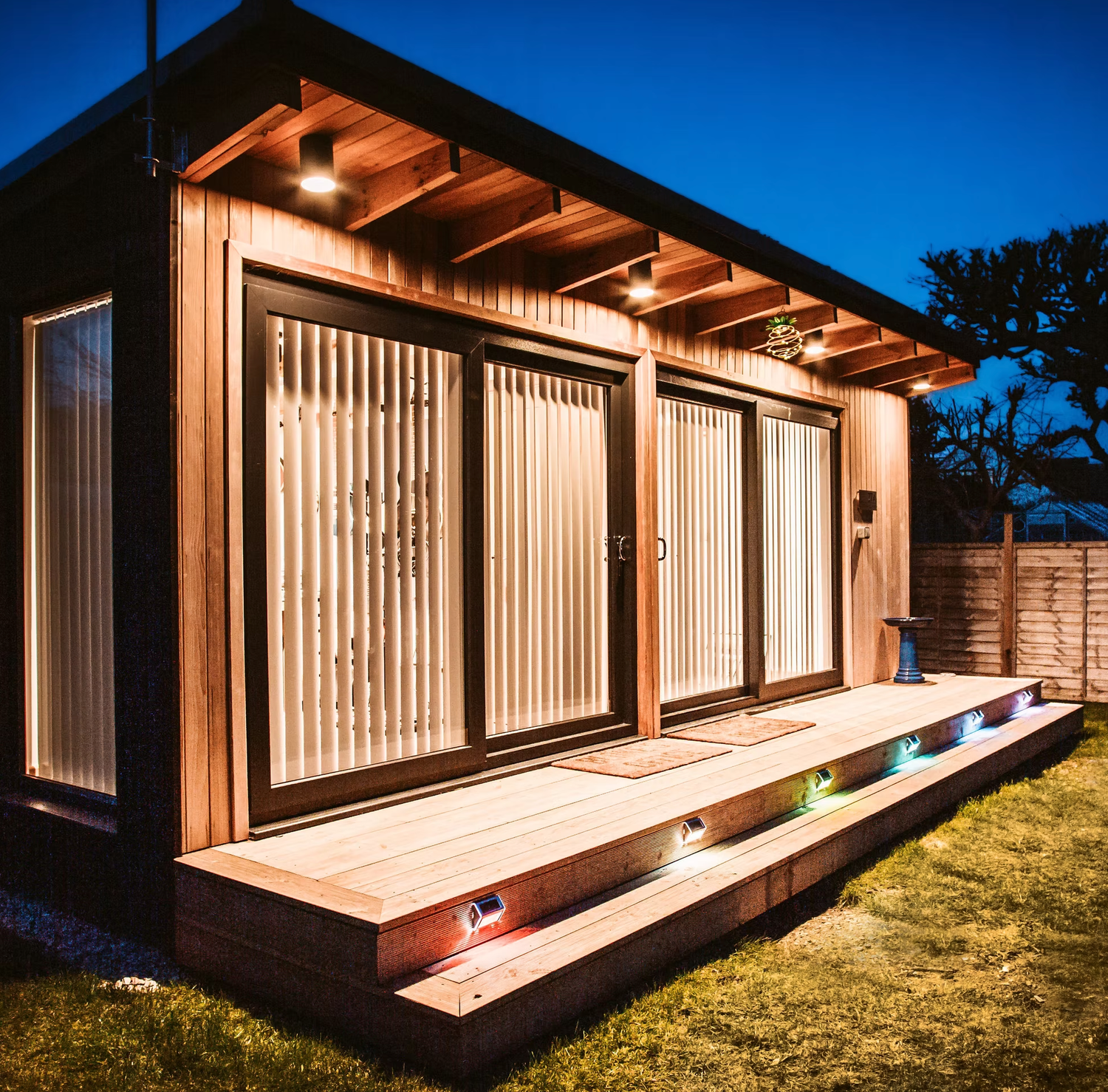 Modern wooden house with large glass windows and deck illuminated at dusk against deep blue sky.