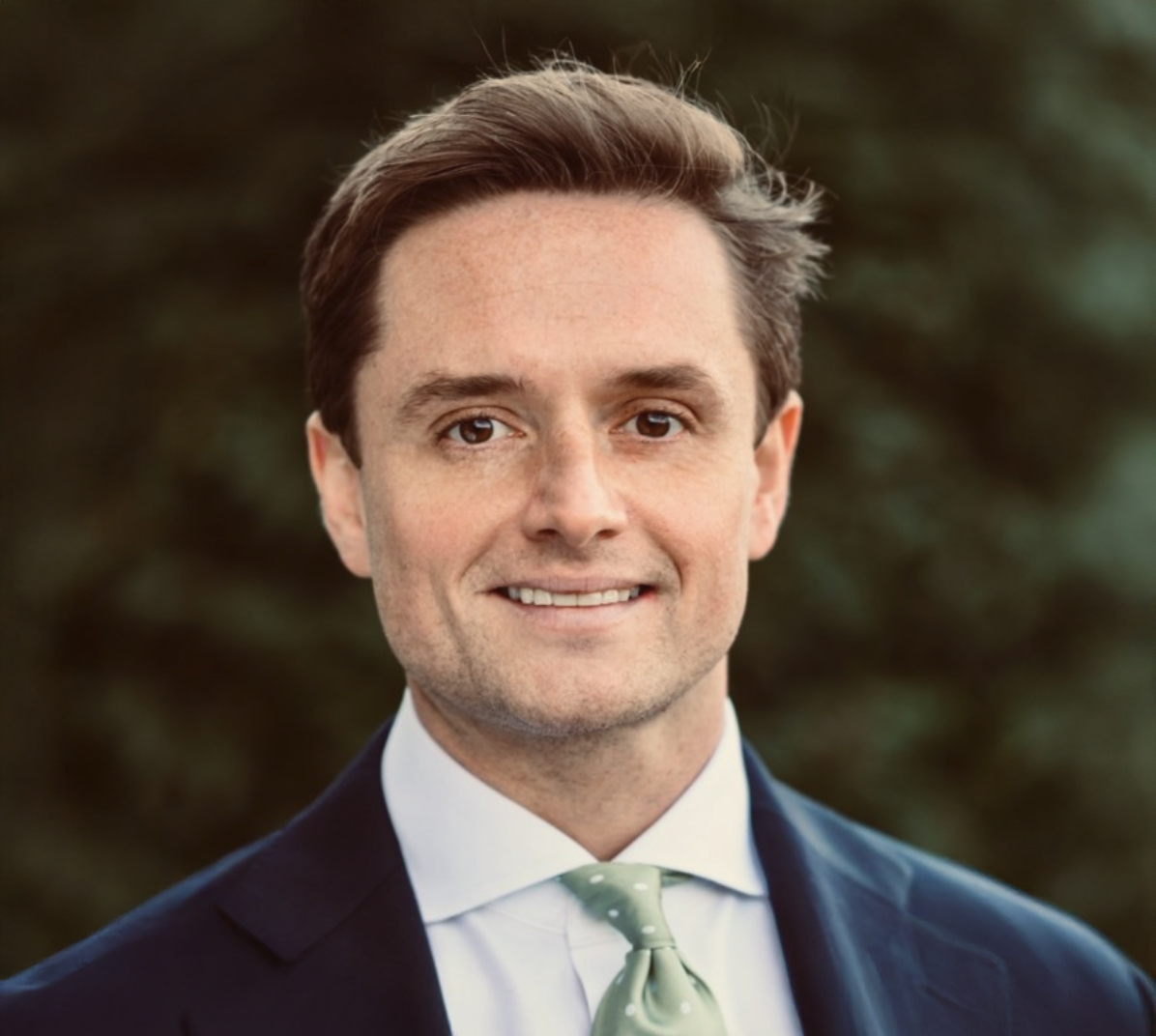 Professional headshot of a smiling businessman in a navy suit and green tie outdoors.