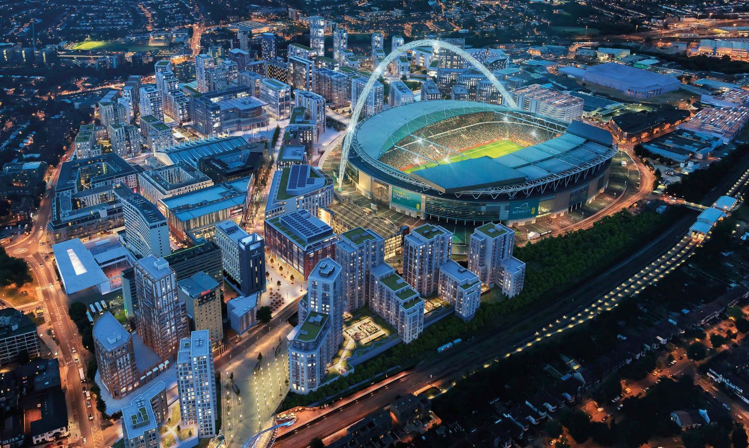 Aerial night view of Wembley Stadium illuminated with its iconic arch, surrounded by urban development in London.