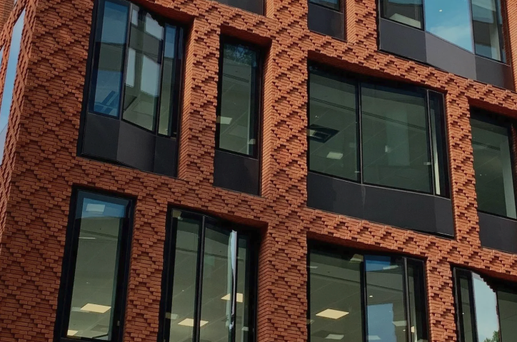 Modern red brick building facade with geometric patterned brickwork and large black-framed windows reflecting natural light.