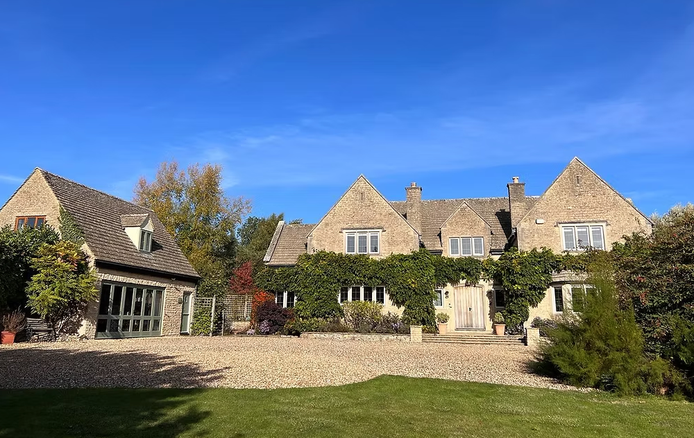 Large stone manor house with gabled dormers and ivy-covered walls under bright blue sky in English countryside setting.