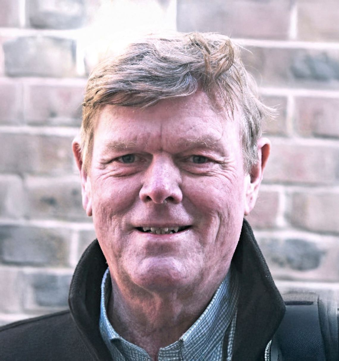 Smiling middle-aged man with gray hair standing in front of a brick wall outdoors.