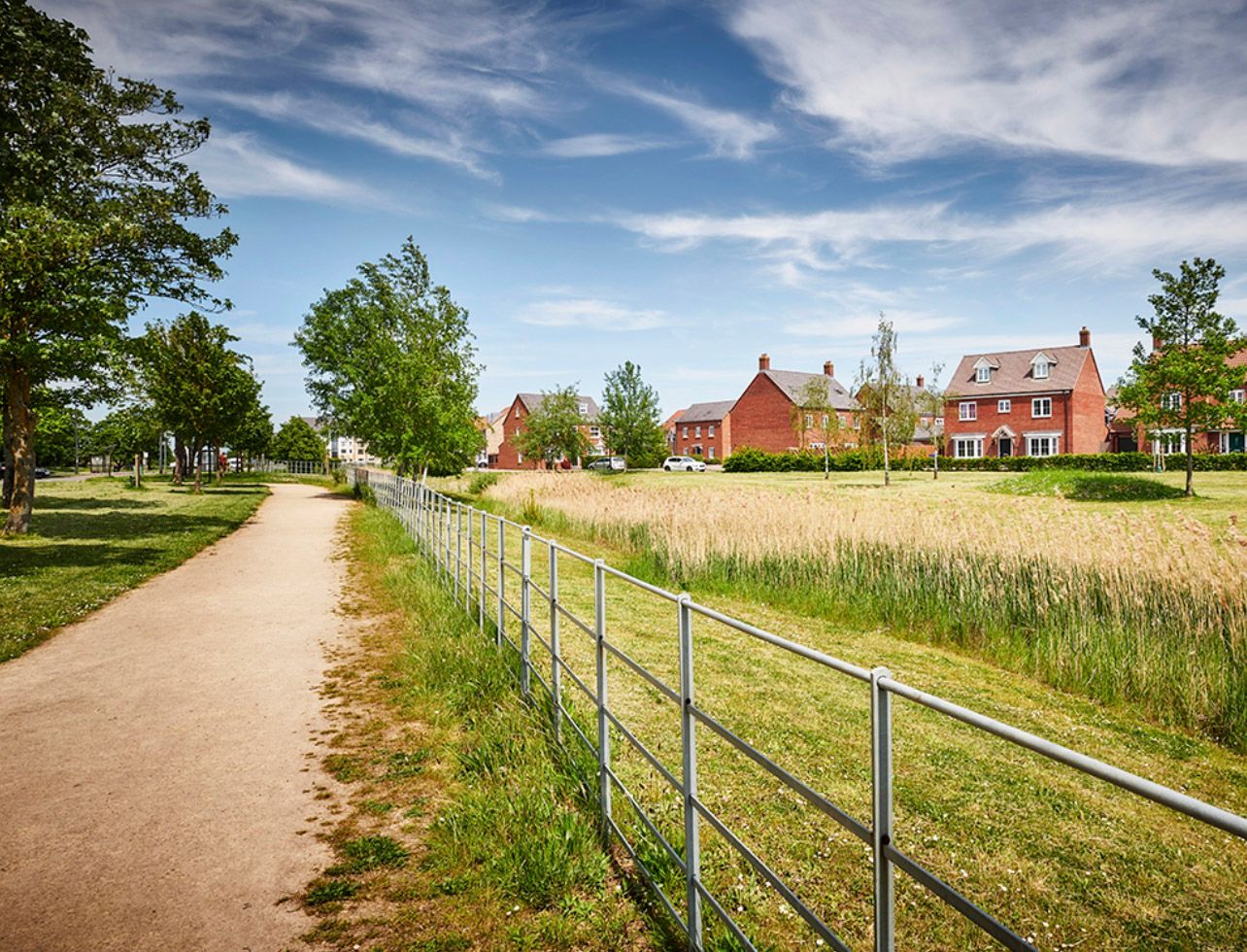 A scenic pathway beside a metal fence leads to red brick houses under a blue sky in a peaceful English suburban neighborhood.