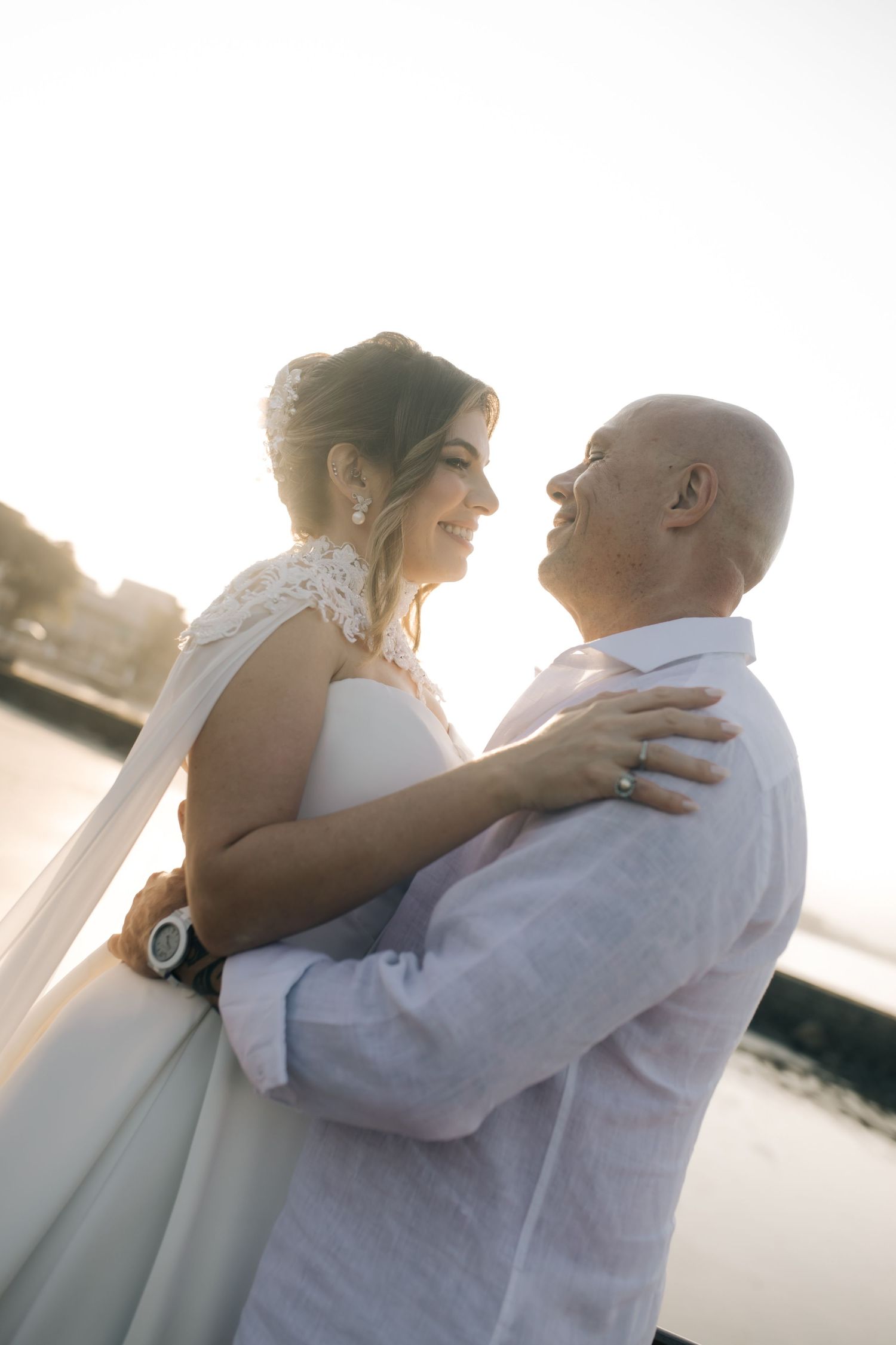A sunset portrait of newlyweds embracing by the water in all-white attire.