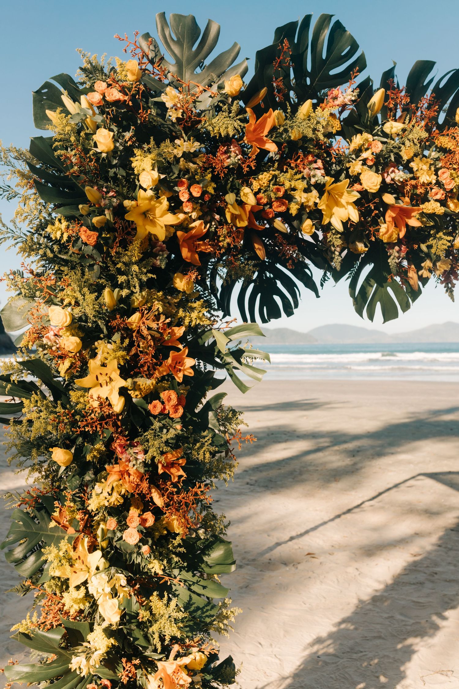 Tropical wedding arch with orange and yellow flowers against ocean backdrop.
