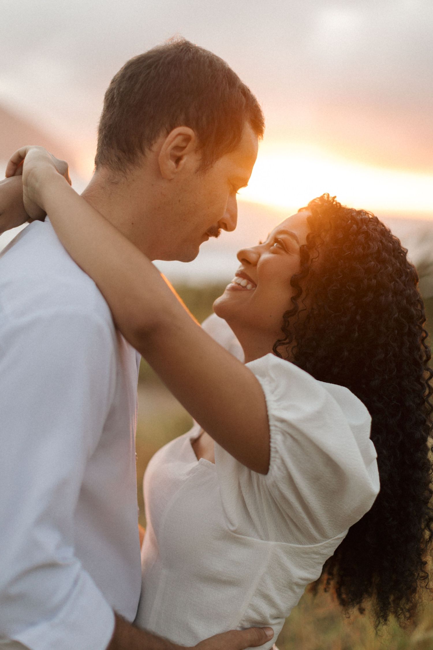 A couple in white outfits share an intimate romantic moment together at sunset, creating a warm silhouetted scene.