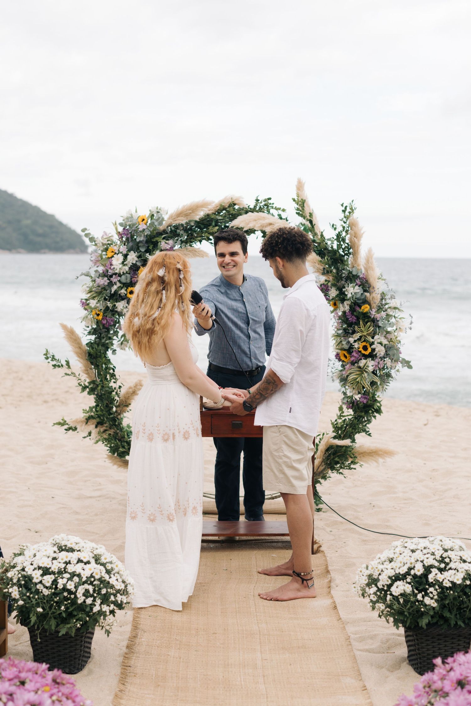 Beach wedding ceremony under a floral arch with mountains in the background and potted flowers lining the aisle.