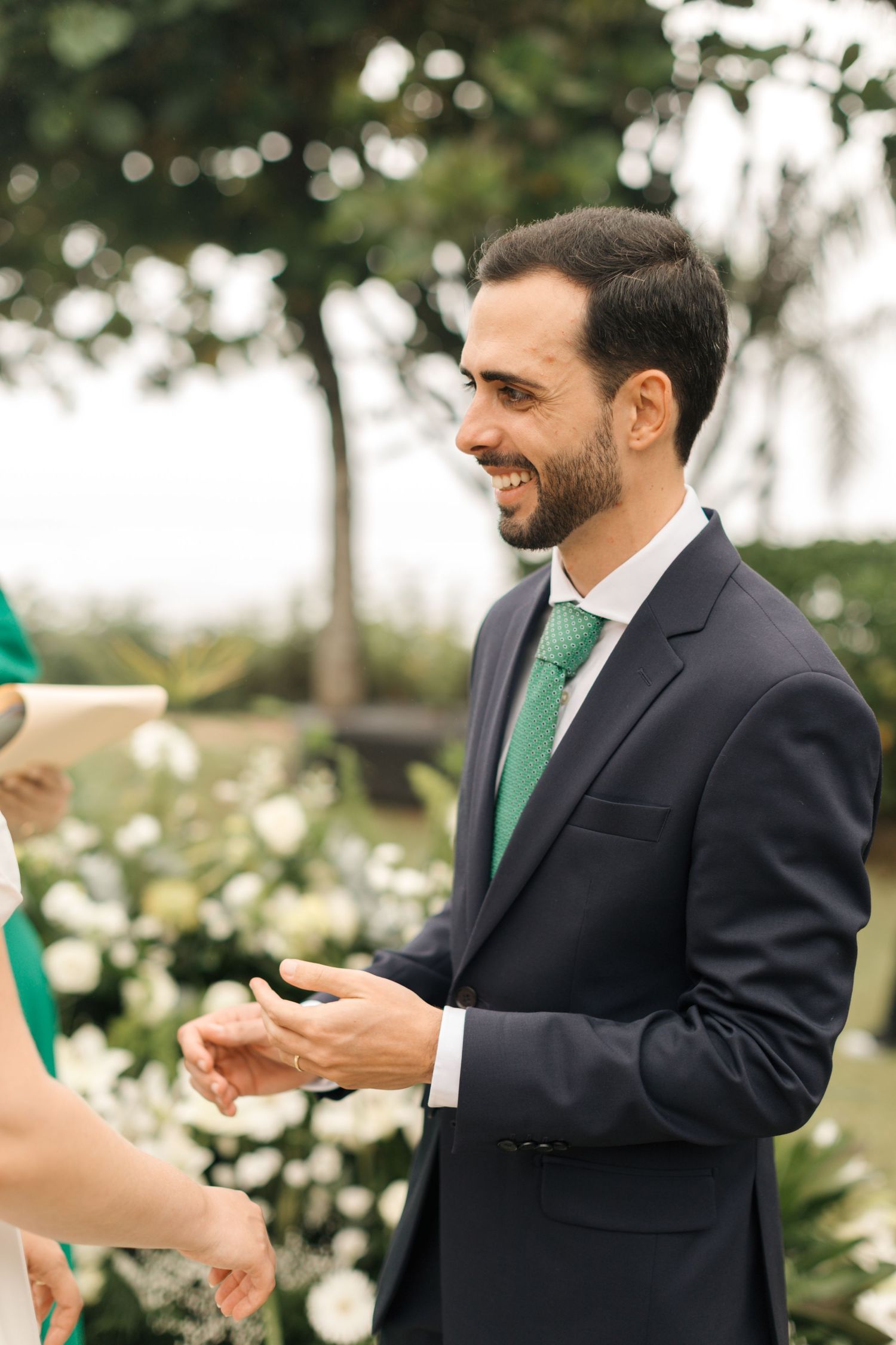 A man in a navy suit with green tie smiles warmly during an outdoor wedding ceremony.
