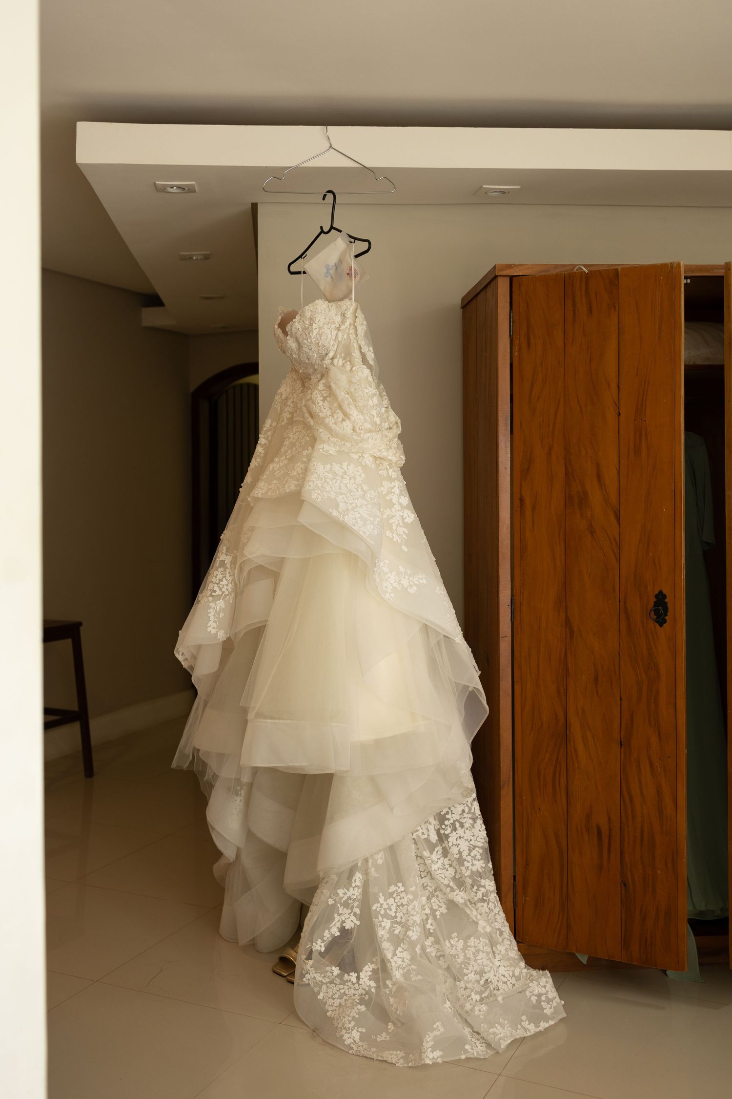 A white lace wedding dress with layered tulle skirt hangs elegantly against a wooden wardrobe.