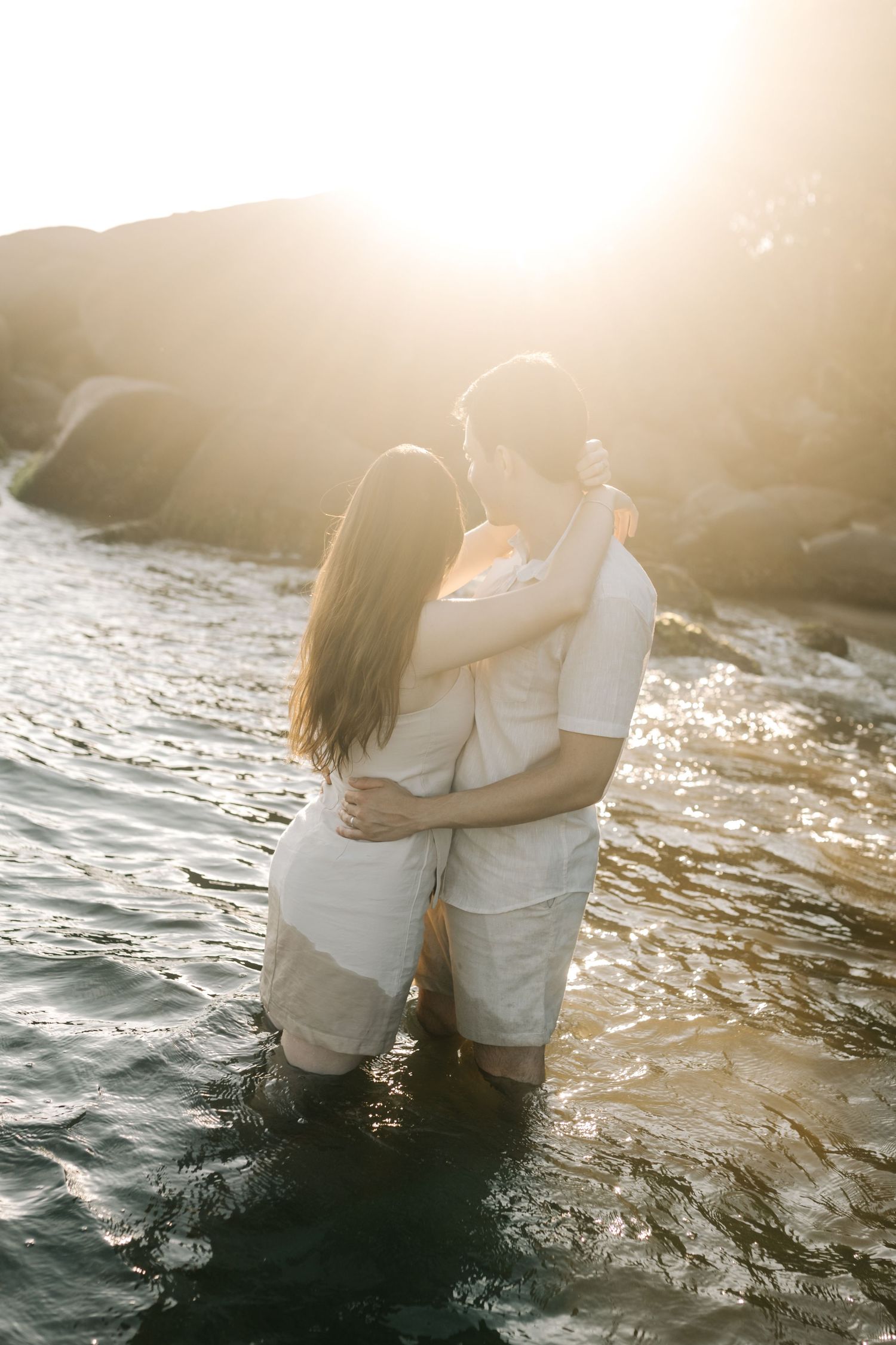 A romantic couple embraces while standing in ocean waters during golden hour.