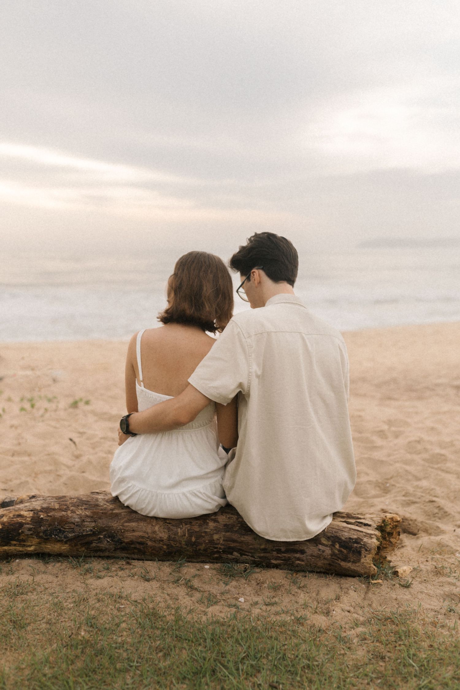 A couple in white clothing sits together on a log overlooking a scenic mountain landscape during sunset.