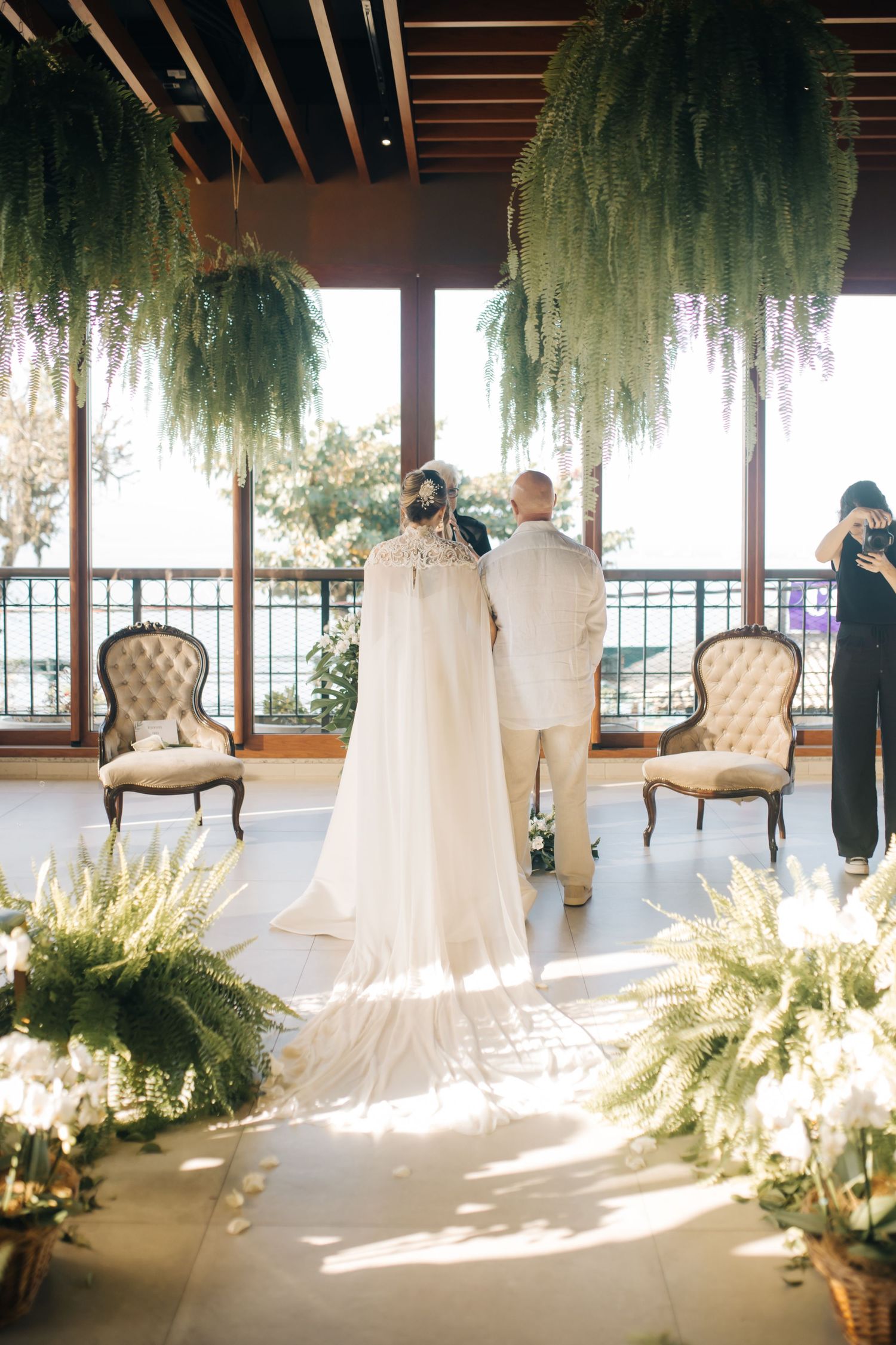 Sunlit wedding venue decorated with hanging ferns and elegant cream-colored chairs.