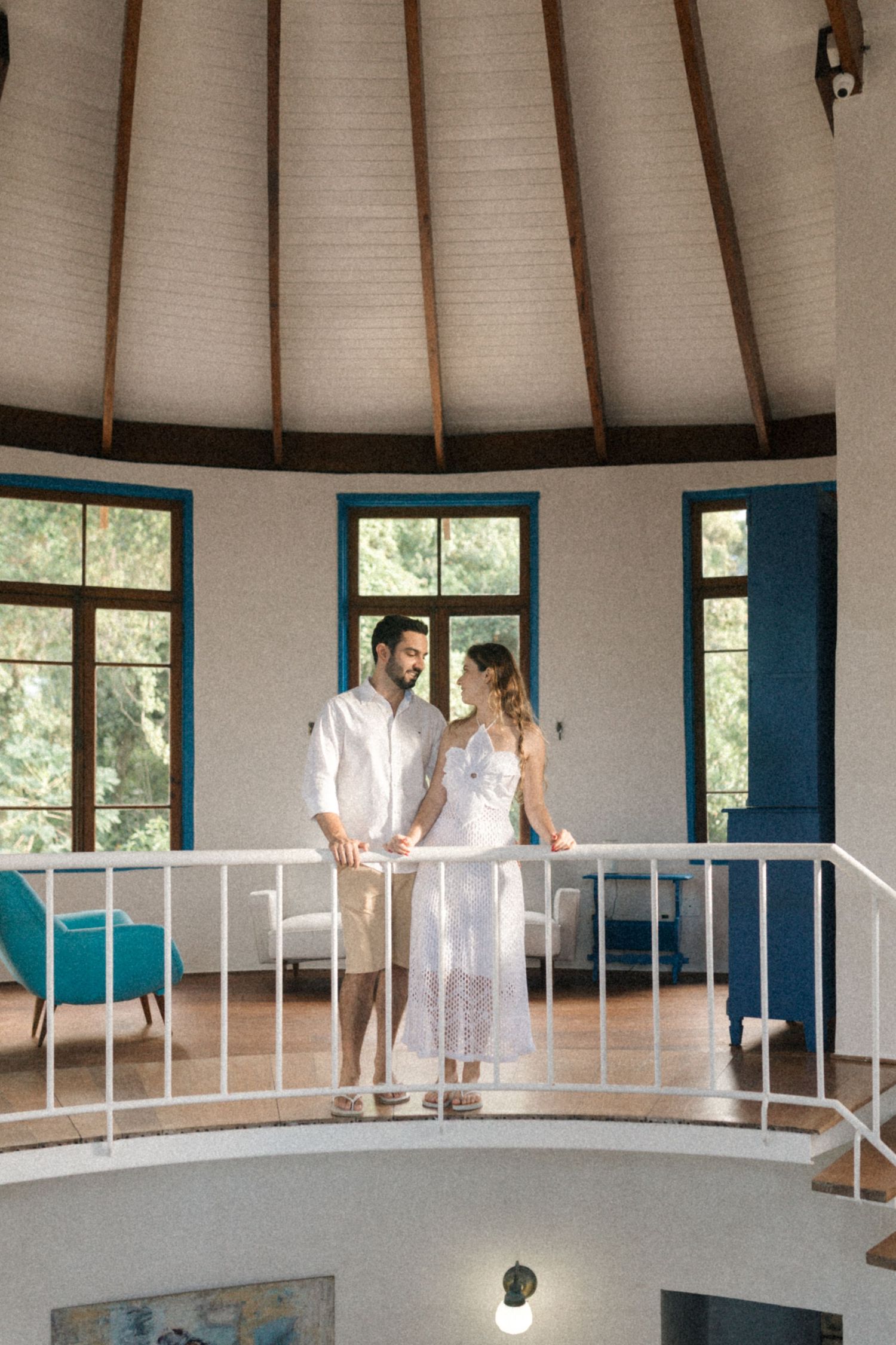 A couple stands together on an indoor balcony surrounded by windows with turquoise trim.