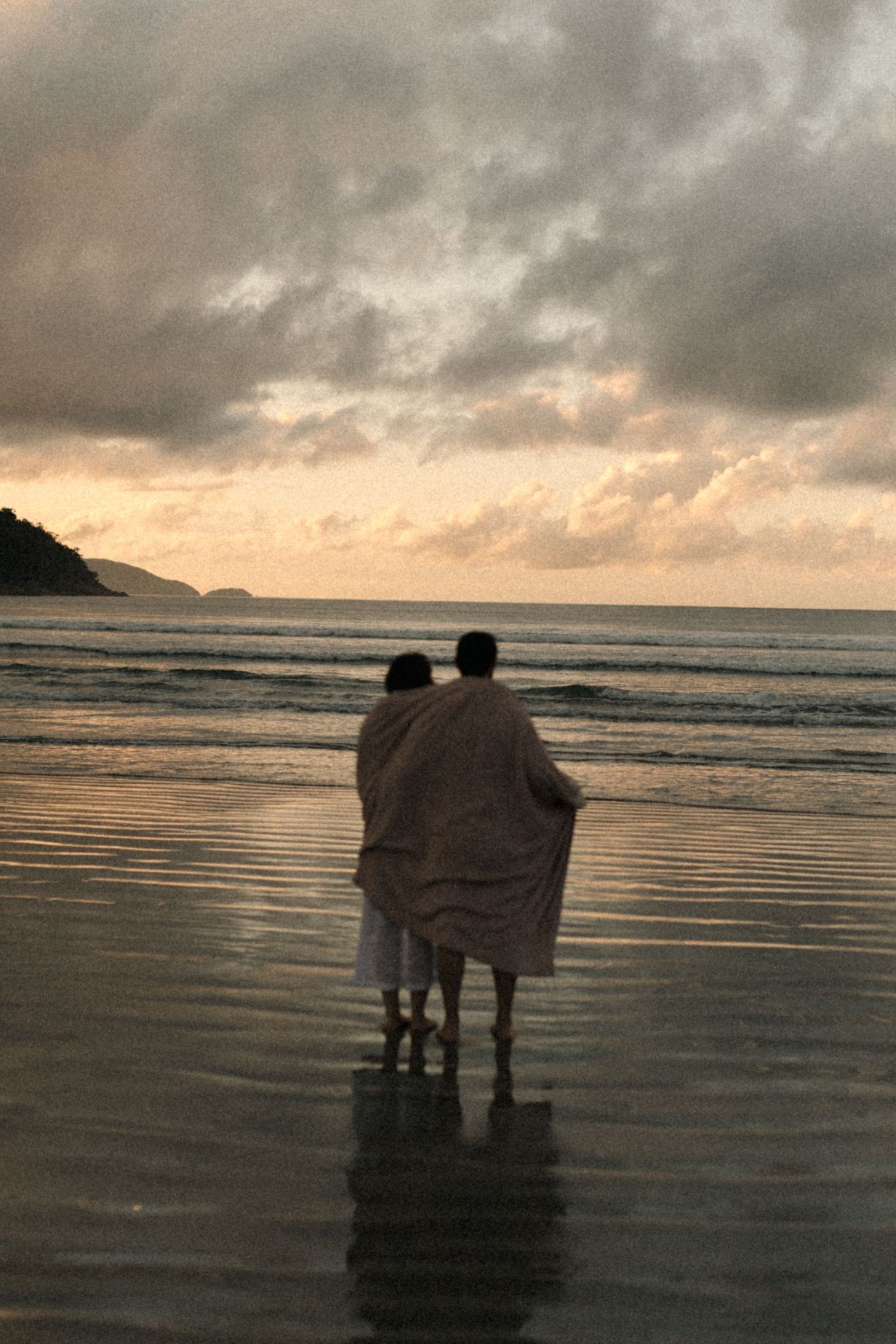 Two figures wrapped in blanket stand on beach during golden hour sunset.