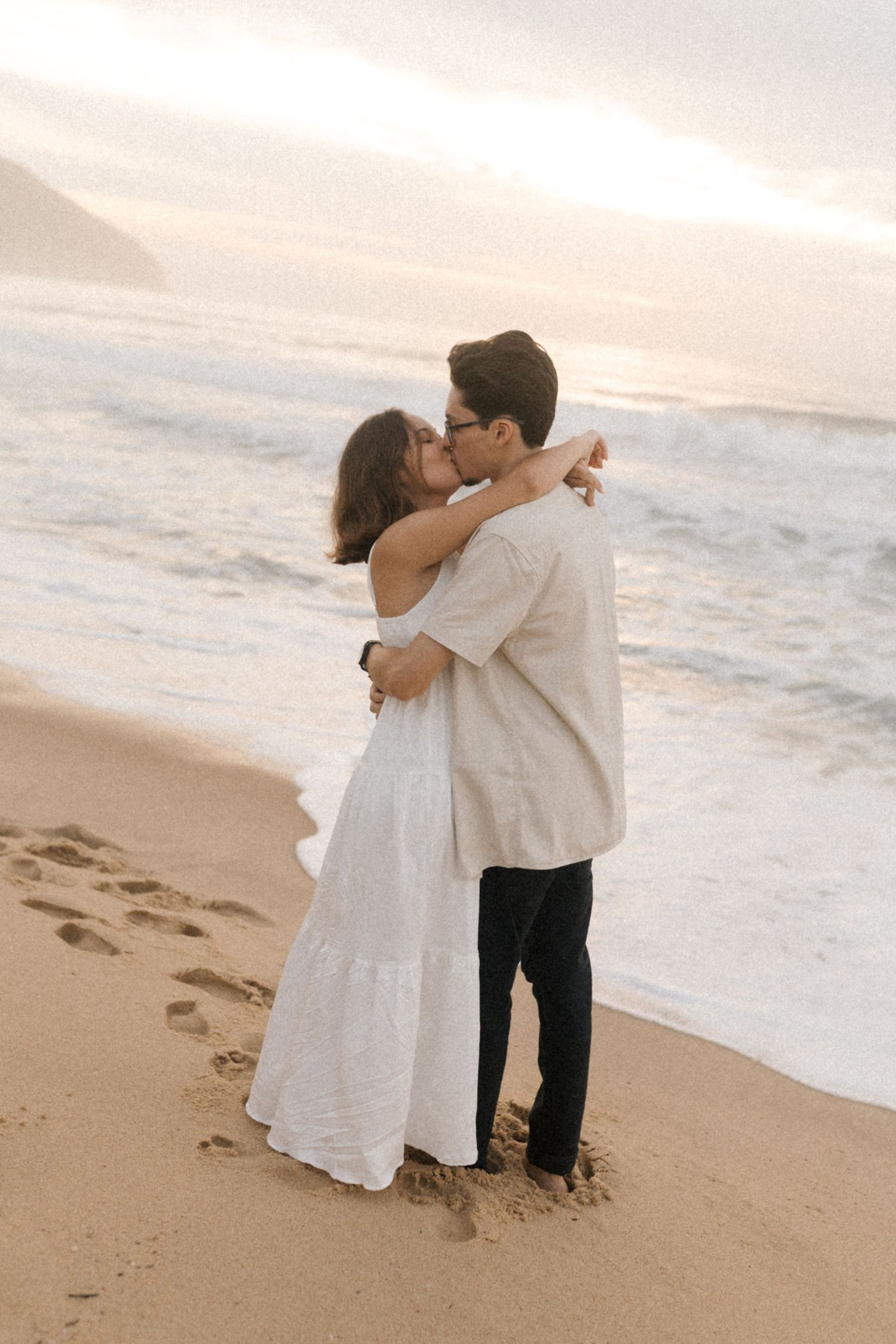 Couple sharing intimate moment on sandy beach at sunset with ocean waves in background.