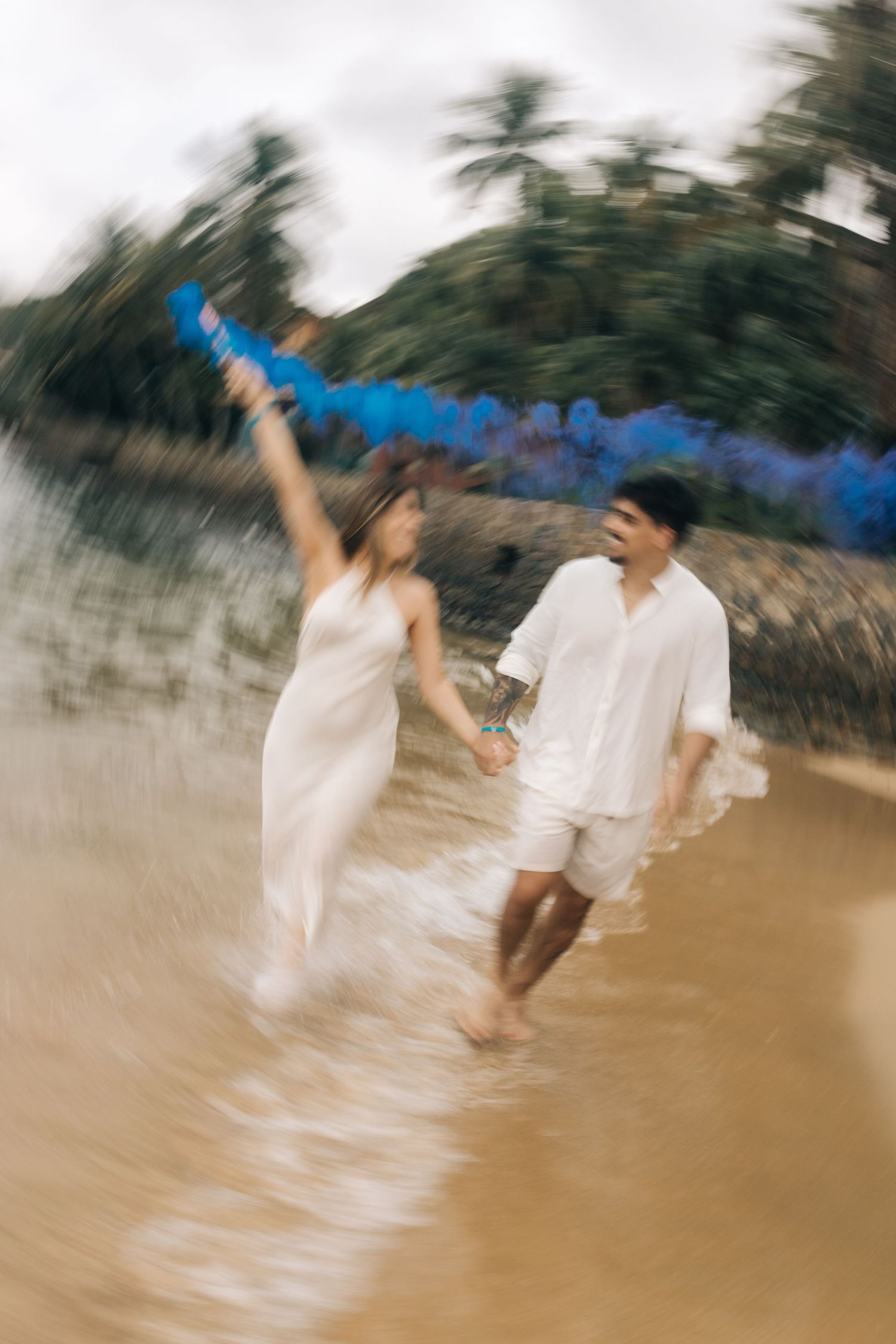 Two people hold hands while splashing through beach water with a bright blue smoke trail above them.