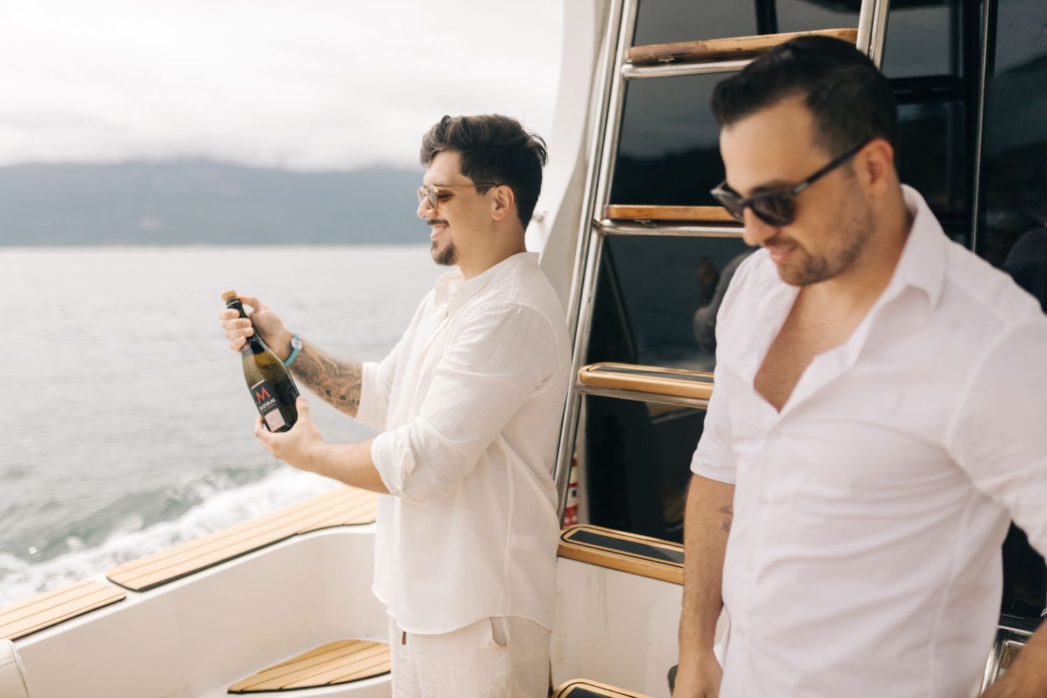 Two friends in white shirts enjoying drinks on a boat deck with ocean views.