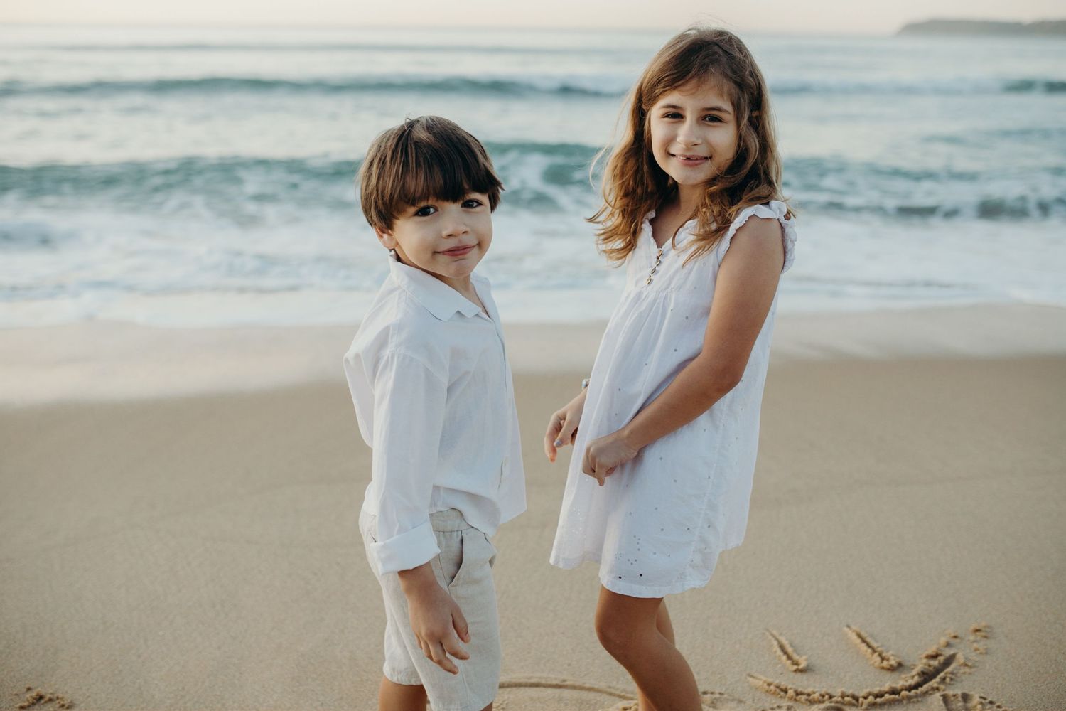 Two children in white outfits playing joyfully together on a scenic beach at sunset.