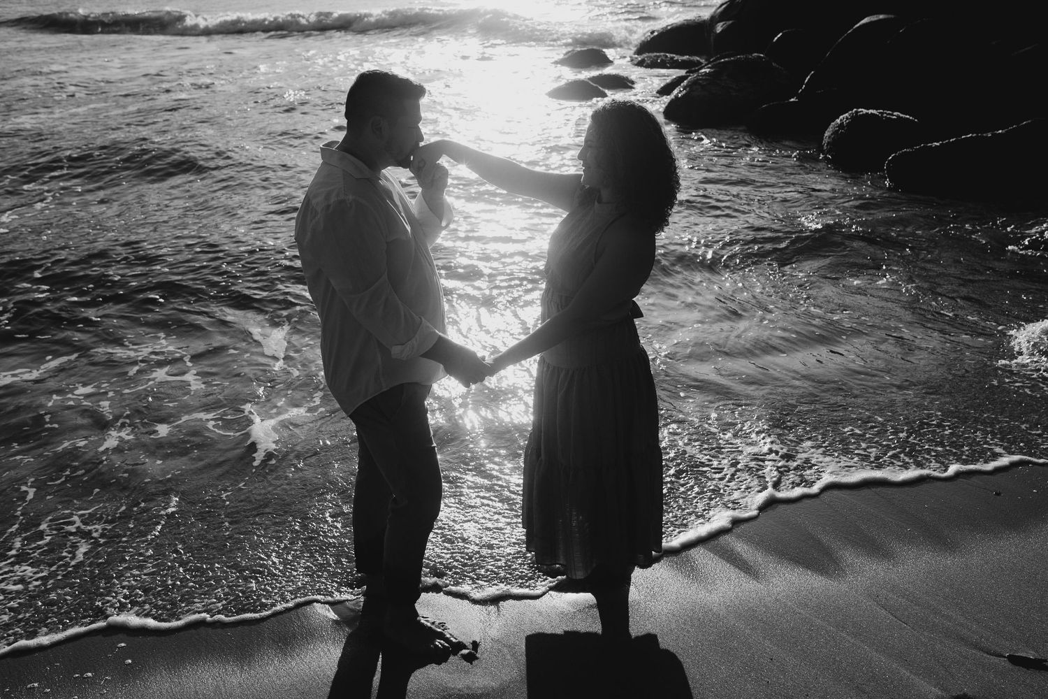 Silhouetted couple holding hands on beach at sunset with waves crashing behind them.