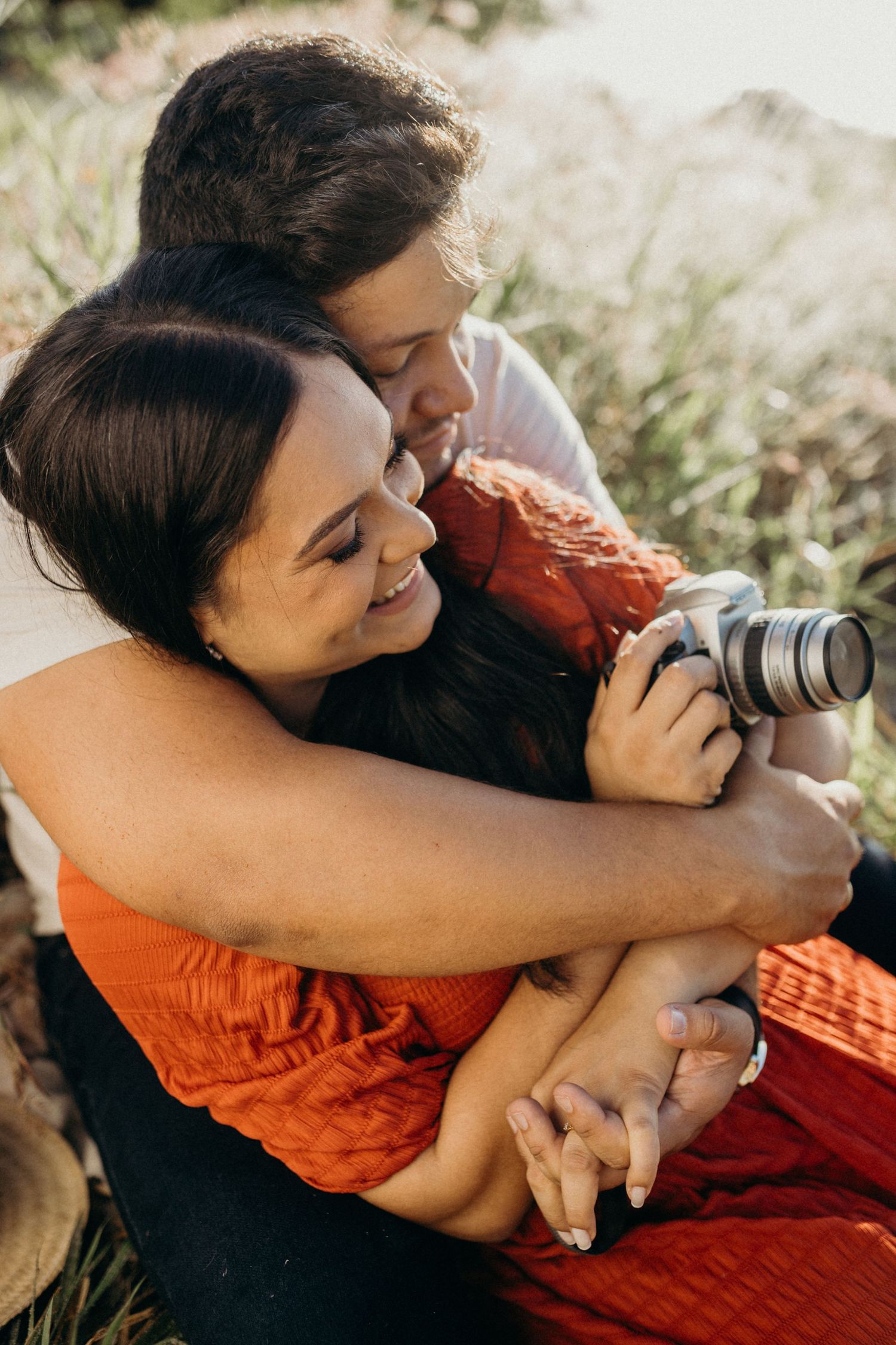 A romantic embrace between two people sharing a camera in sun-drenched coastal grassland.