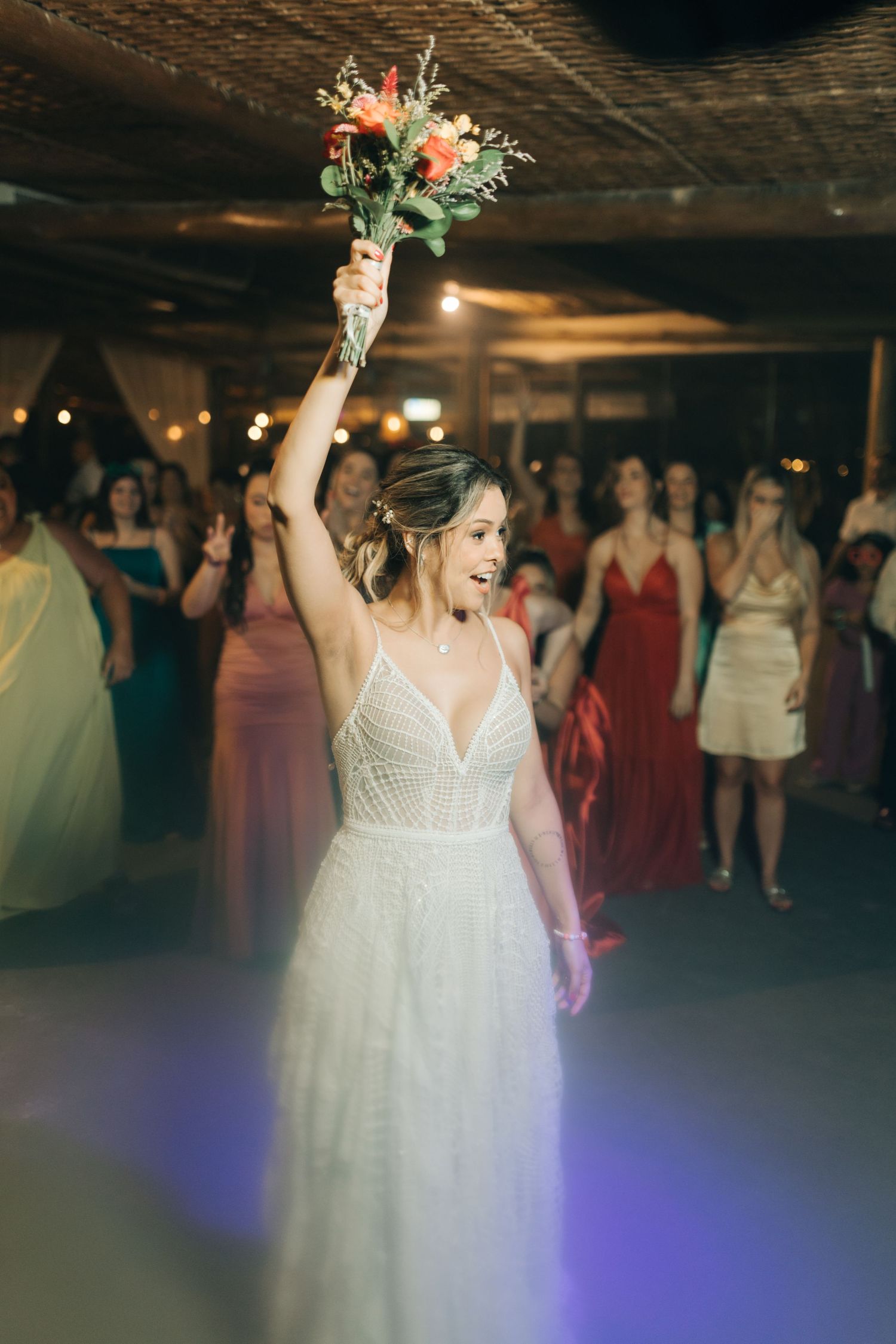 A bride in a white dress holds up a bouquet while dancing at a wedding reception with guests in the background.