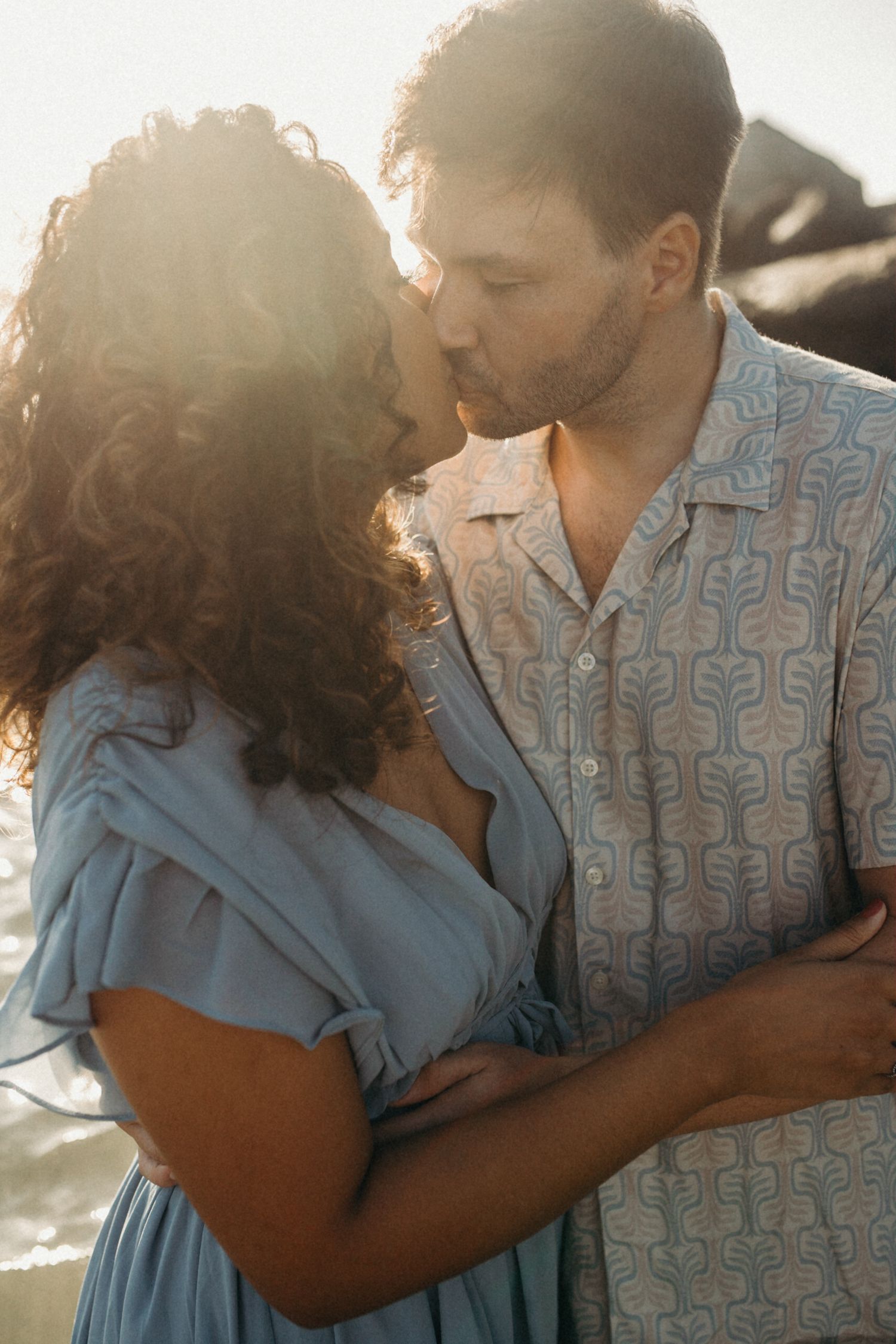 Intimate couple moment captured in warm sunlight on coastal rocks with ocean waves.